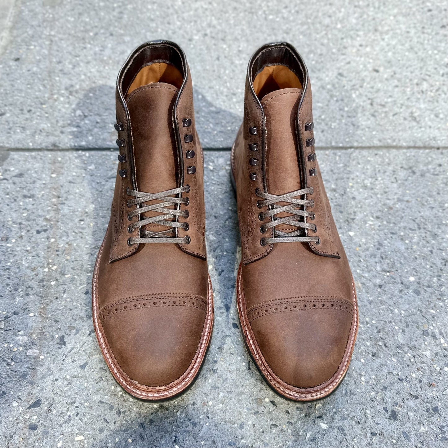 A pair of Alden Madison D2817HC - Straight Tip Shaking Boots in Tobacco Smooth Chamois with brogue detailing and a Commando Sole rests on a speckled gray pavement, showcasing a slightly worn texture for a rustic look.