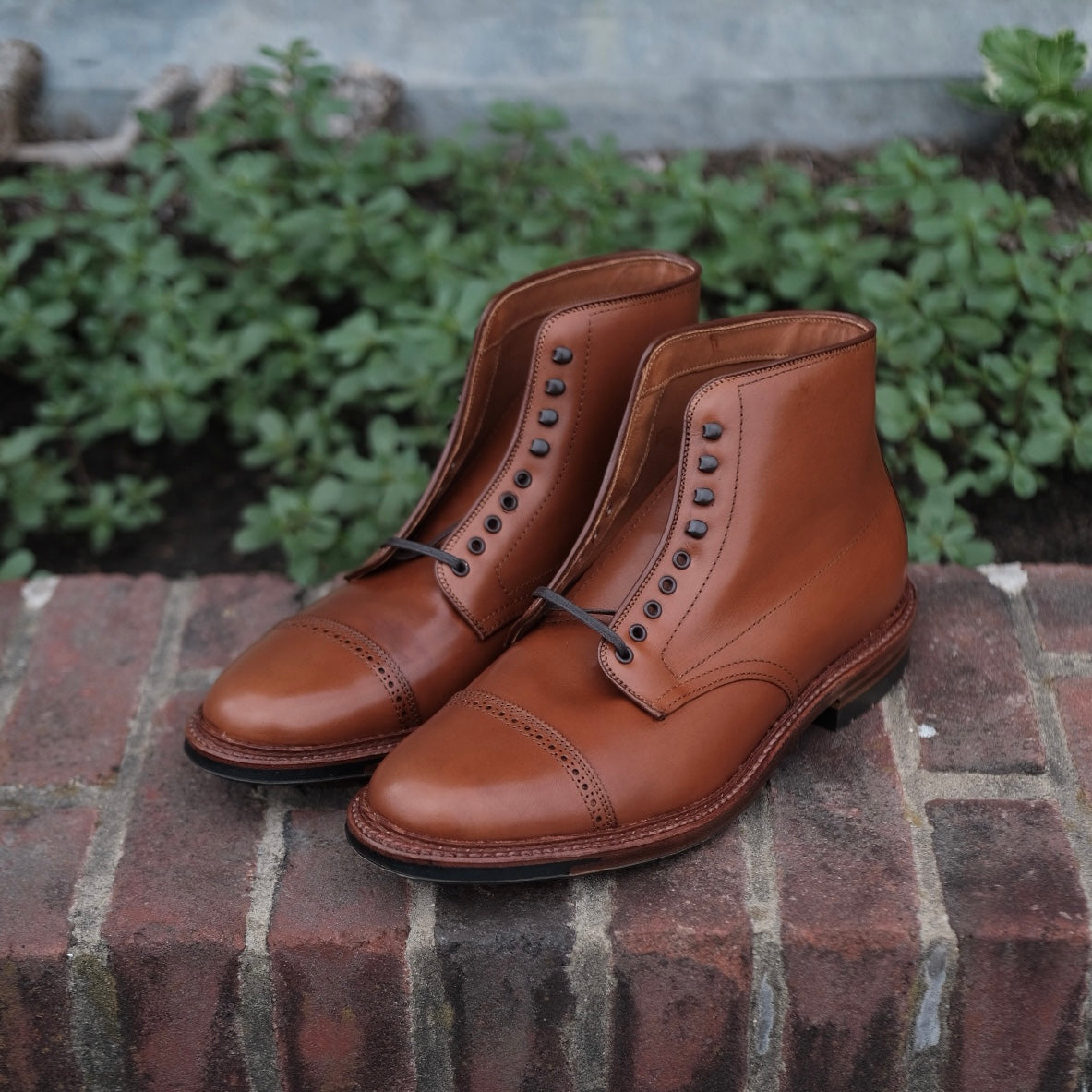 The Alden Madison D2825HC Jumper Boot in Burnished Tan Calfskin boasts a sleek design with black eyelets and laces on a Barrie Last. Impeccably crafted with visible stitching on the toe cap and commando sole, it sits on a brick surface with blurred green foliage in the background.