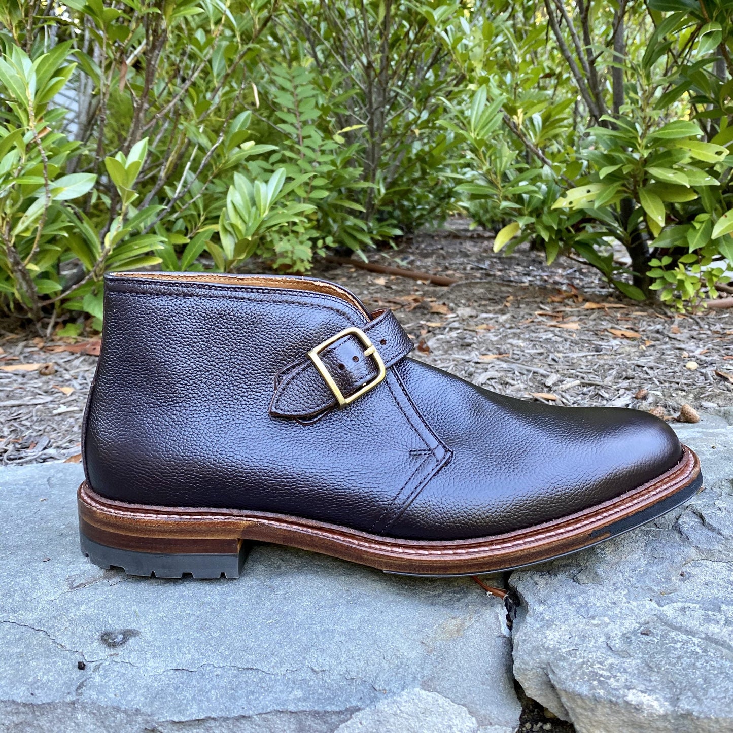 An Alden Madison D9704C - George Boot in Brown Regina Grain, featuring a gold buckle, rests on a stone surface outdoors amidst lush green foliage.