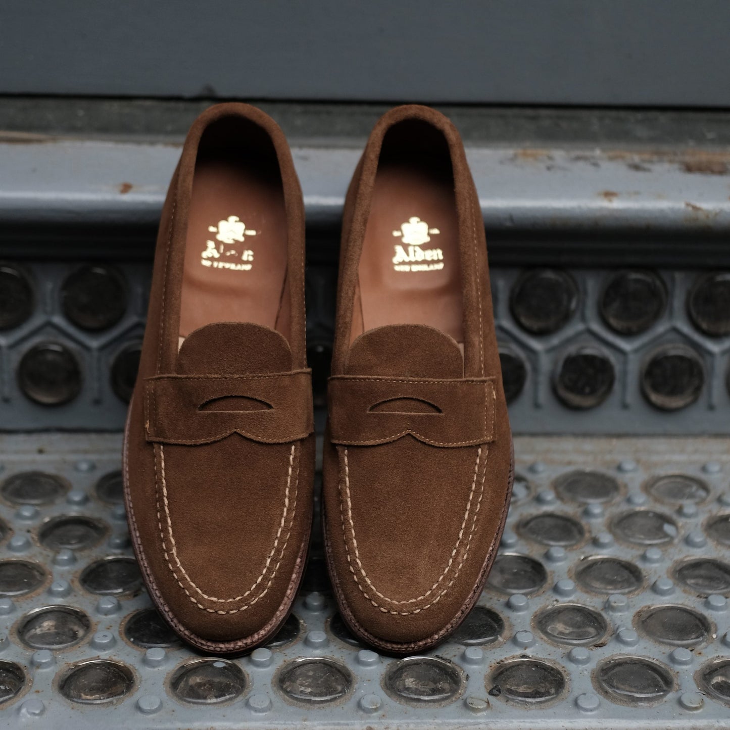 A pair of Alden Madison 6243F - Unlined Leisure Hand Sewn loafers in snuff suede, featuring hand-stitched detailing, rests on a patterned metal surface, showcasing their craftsmanship.