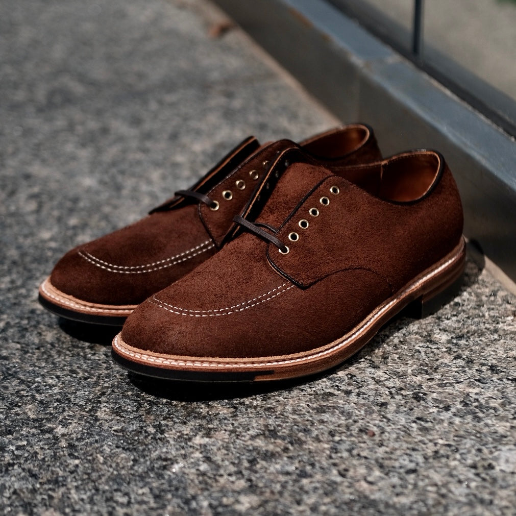 The Alden Madison D2604C - Indy Shoe in Tobacco Reverse Chamois features white stitching and dark laces through metal eyelets. Displayed on a textured gray stone surface, its elegant craftsmanship is effortlessly highlighted beside a reflective glass pane.