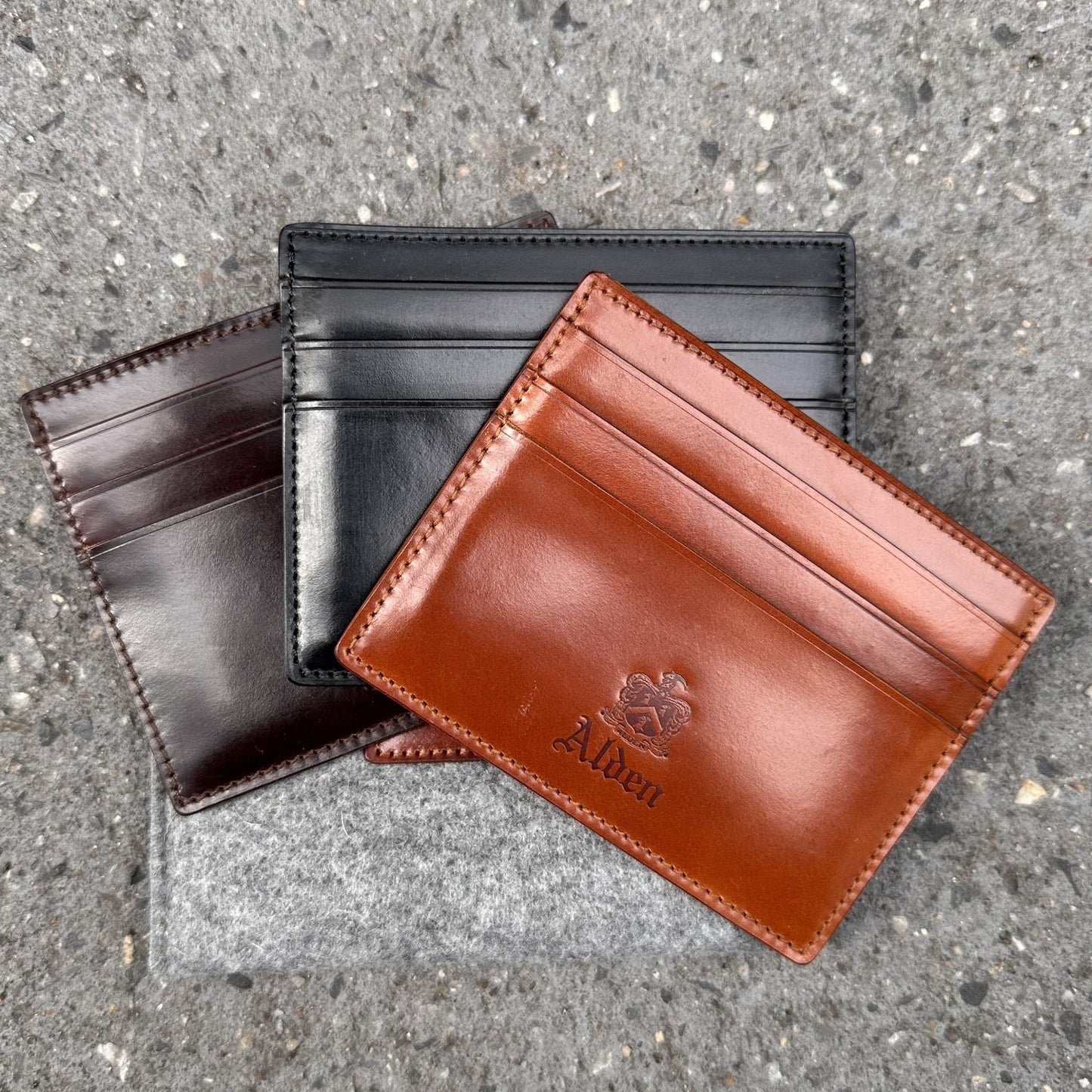 Displayed on a gray, textured surface are three Alden Madison LG 6009 Shell Cordovan card holders in black, dark brown, and brown. The brown holder features an embossed logo and name, with each holder offering six pockets for cards.