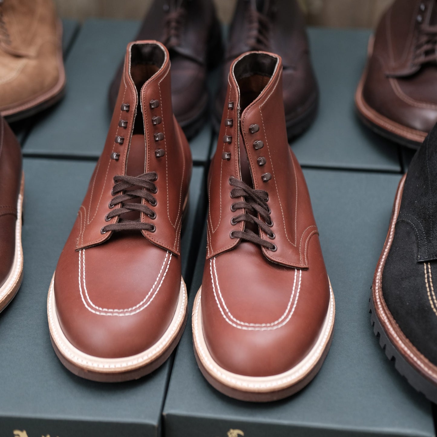 Displayed on a dark green box, the Alden Madison 405 - Indy Boot (Original) in Mid Brown Calfskin features precise eyelets and hooks for laces. In the background, other brown and black boots echo the classic Alden Indy Boot style.