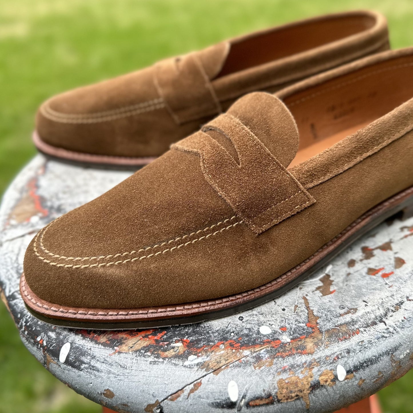 The Alden Madison 6221L Unlined Penny Mocc in Snuff Suede, featuring a rubber sole, is placed on a weathered round table with colorful chipped paint. The blurred background of green grass hints at an outdoor setting.