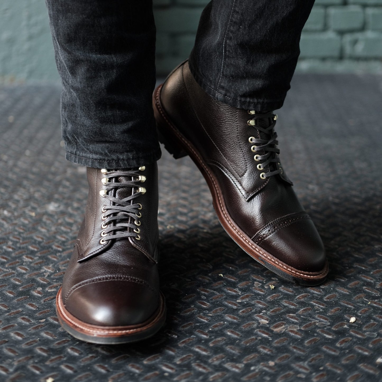 Wearing Alden Madison’s D9819HC Jumper Boot in Regina Brown Grain, a person pairs dark leather lace-up boots with black pants, standing on a textured metal surface against a greenish-gray brick wall.