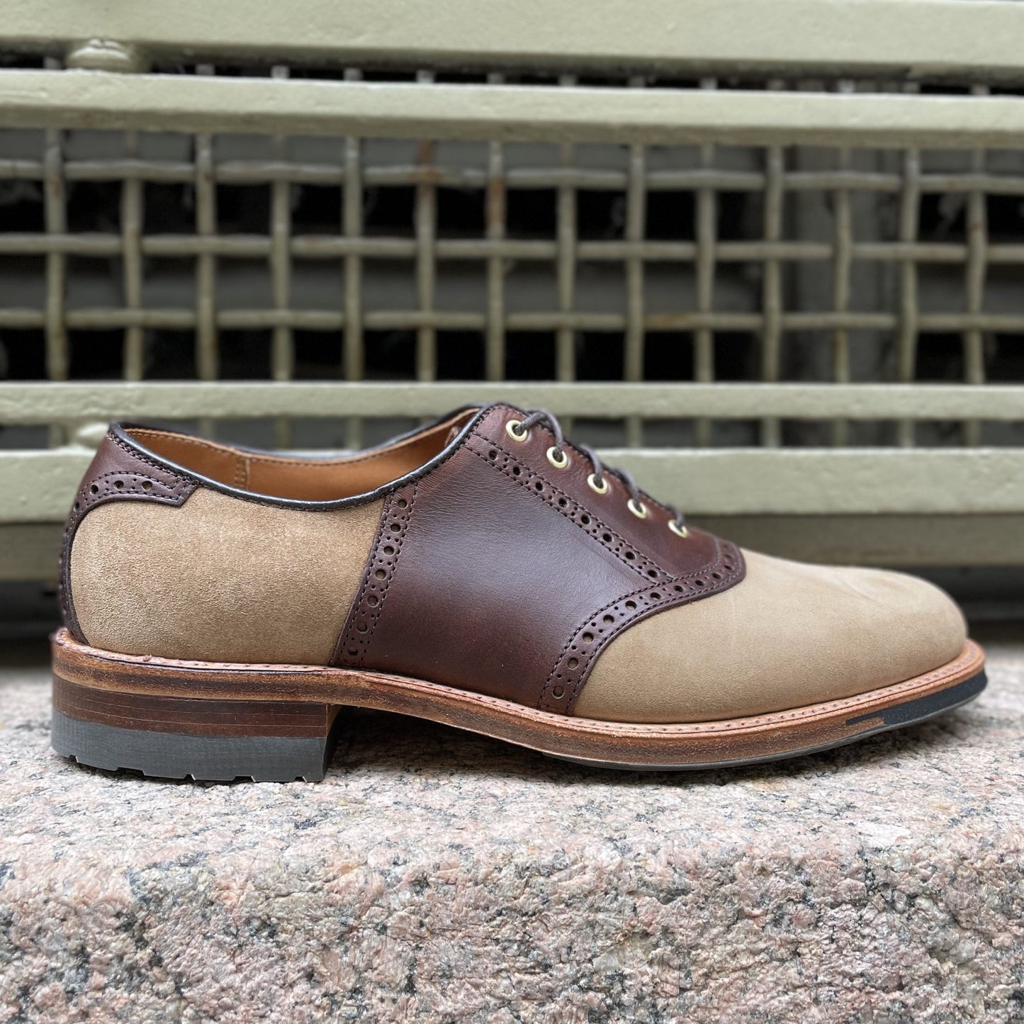 The Alden Madison Valhalla Saddle Oxford, in CXL and tan suede with brogue detailing and a robust commando sole, sits stylishly on a stone surface against a metal grate backdrop.