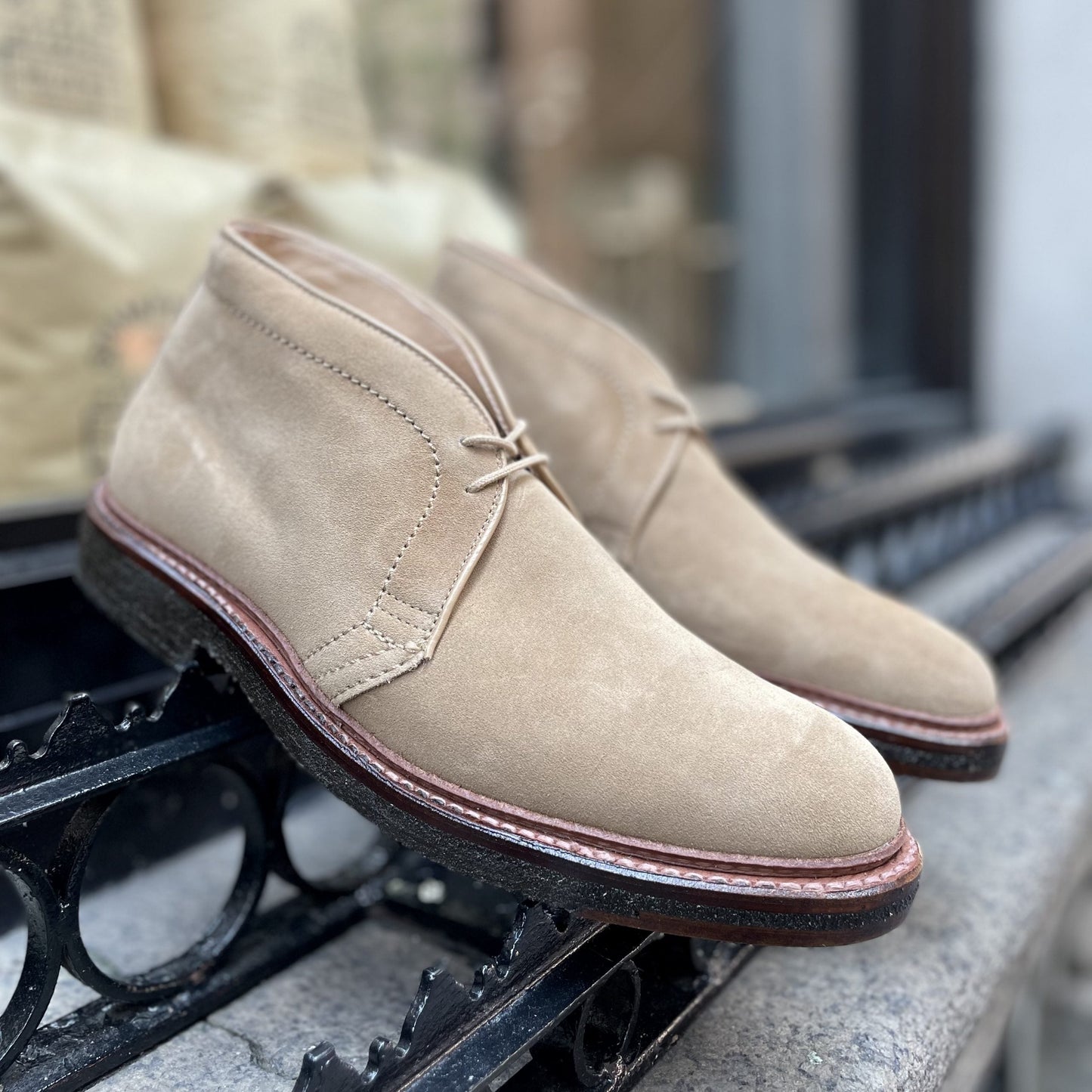 A pair of Alden Madisons 14580 Desert Chukka Boots in Tan Suede, elegantly showcased on a black wrought iron railing outdoors, features a sleek design with lace-up fronts, brown soles, and visible edge stitching that enhances their timeless appeal.