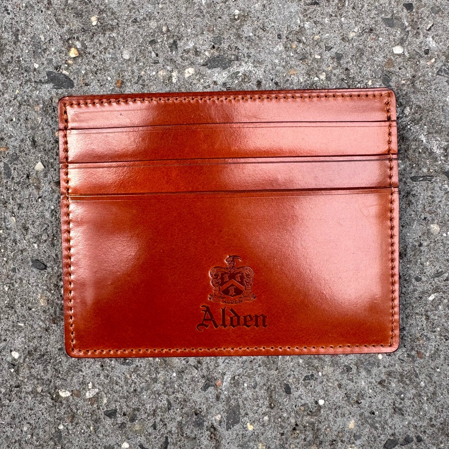 The Alden Madison LG 6005 Cognac Credit Card Holder is crafted from luxurious Shell Cordovan leather, embossed with Alden and a crest. Set on a textured gray surface, it features six card pockets, merging functionality with timeless style inspired by Alden Shoe artistry.
