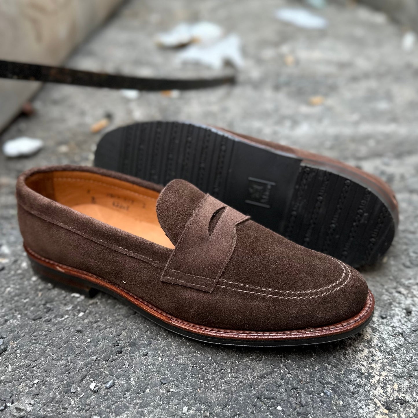 Alden Madisons 6224L Unlined Penny Mocc in brown suede, featuring mocc toe and penny slot detail with a smooth leather lining and black rubber soles, is displayed on textured concrete with one shoe resting on its side.