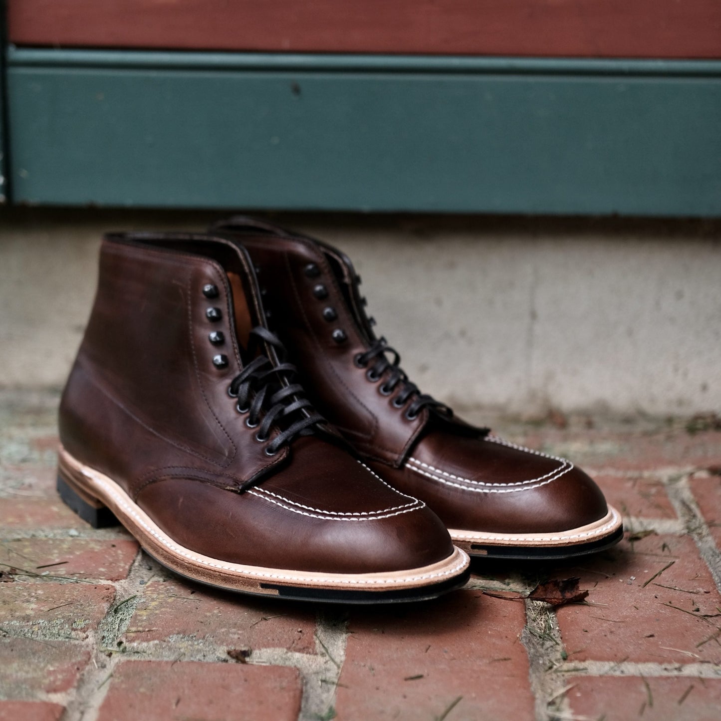 The dark brown 403C Indy Boot with Commando Sole in CXL by Alden Madison features black laces, white toe stitching, and sits on a brick surface against a green-red backdrop.
