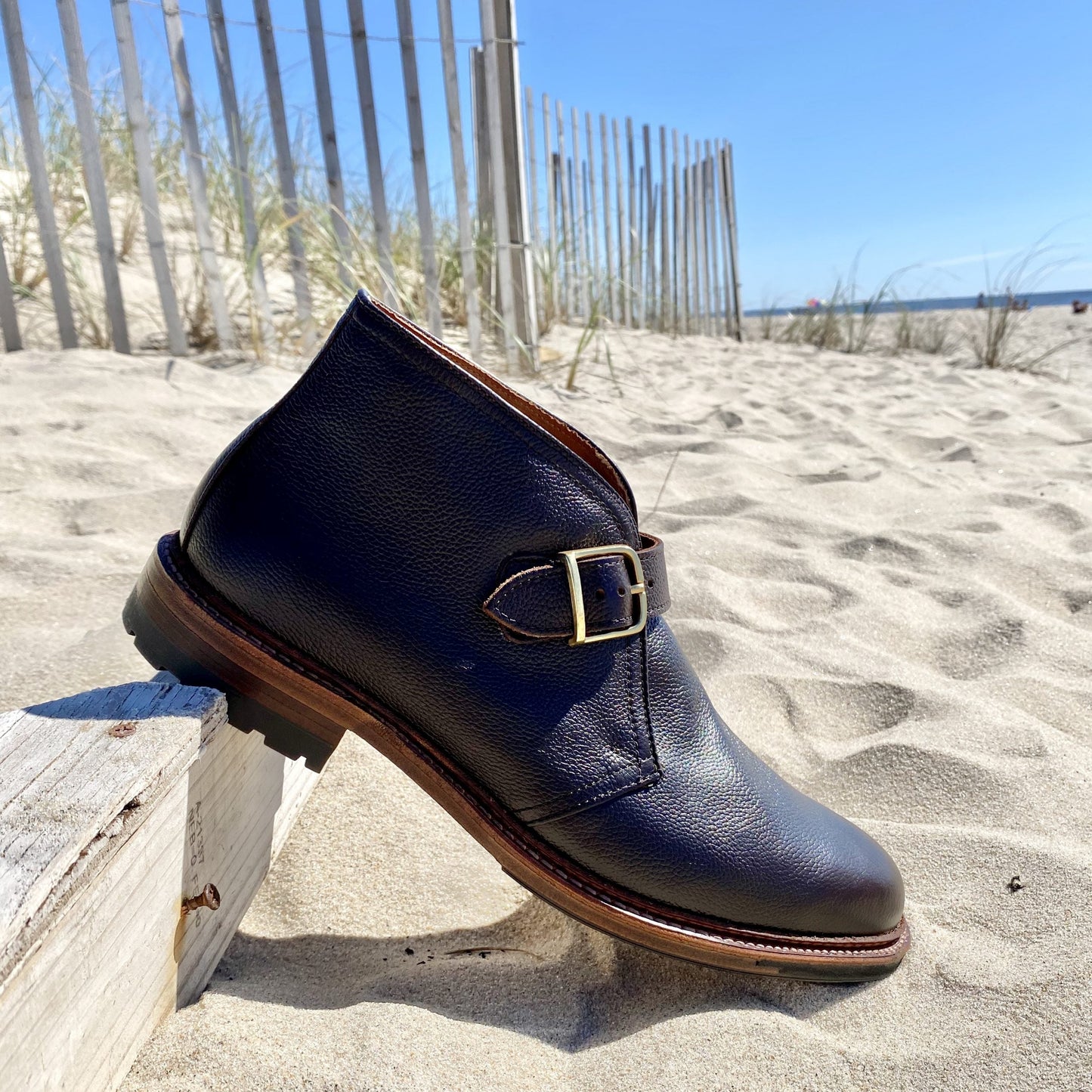 A single Alden Madison D9704C - George Boot in Brown Regina Grain, featuring dark brown leather and a gold buckle, rests on sand near a wooden fence at the beach under a clear blue sky.