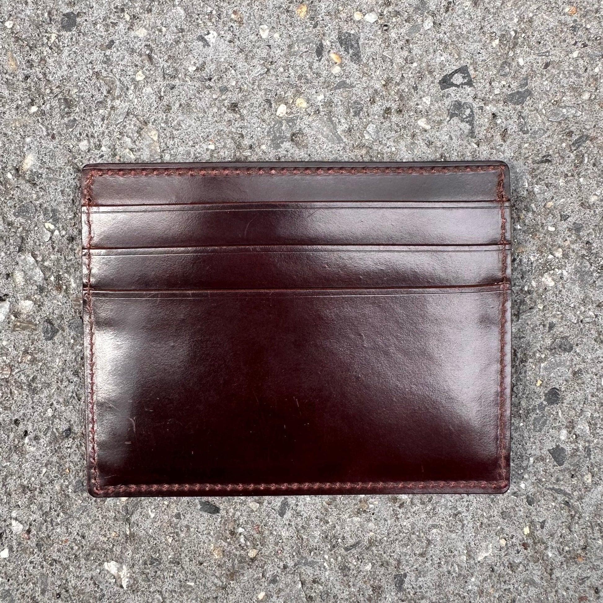 On a textured gray concrete surface lies the LG 6008 - Alden Color 8 Shell Credit Card Holder, a dark brown leather creation by Alden Madison. Featuring three visible slots, its crafted from premium Shell Cordovan with a smooth, polished finish reminiscent of an Alden Shoes elegance.