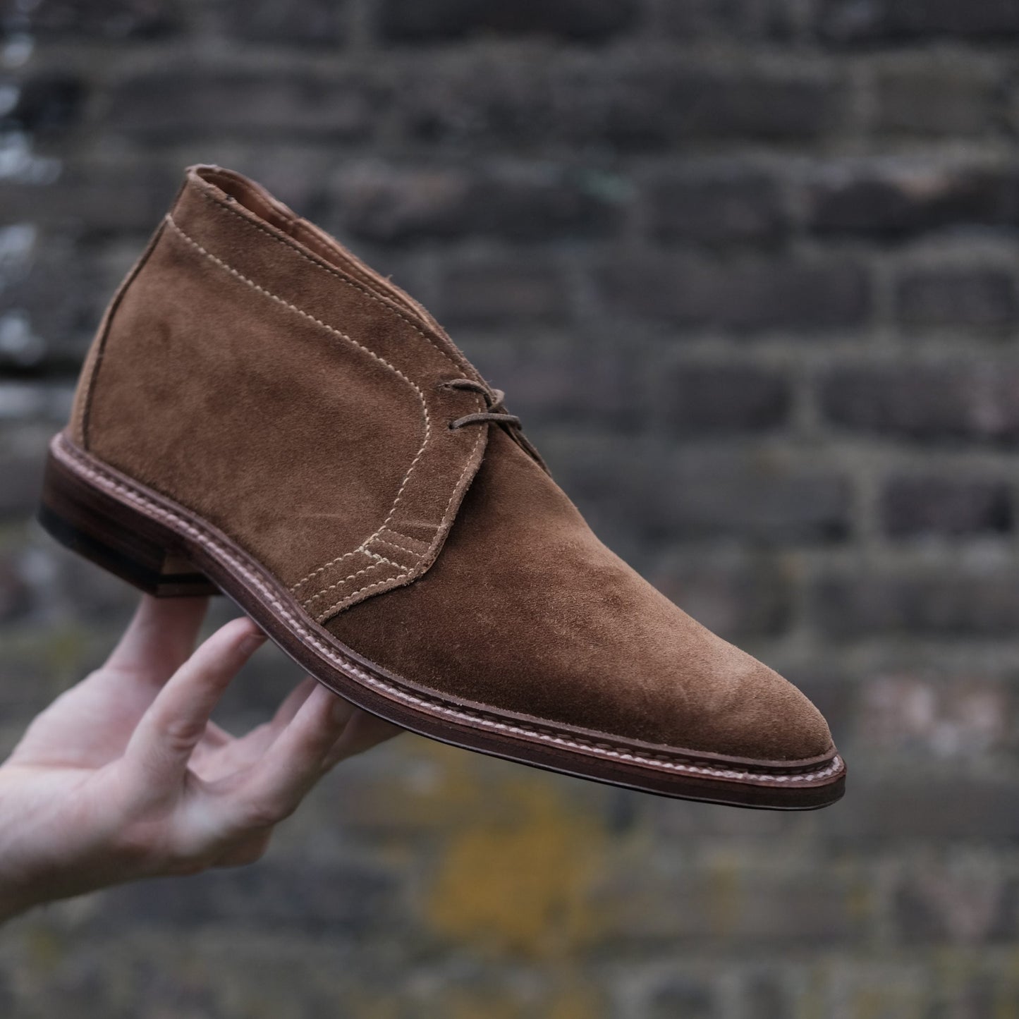 A hand cradles the Alden Madison 1493 - Unlined Chukka in Snuff Suede, with visible stitching glinting against a weathered brick wall.