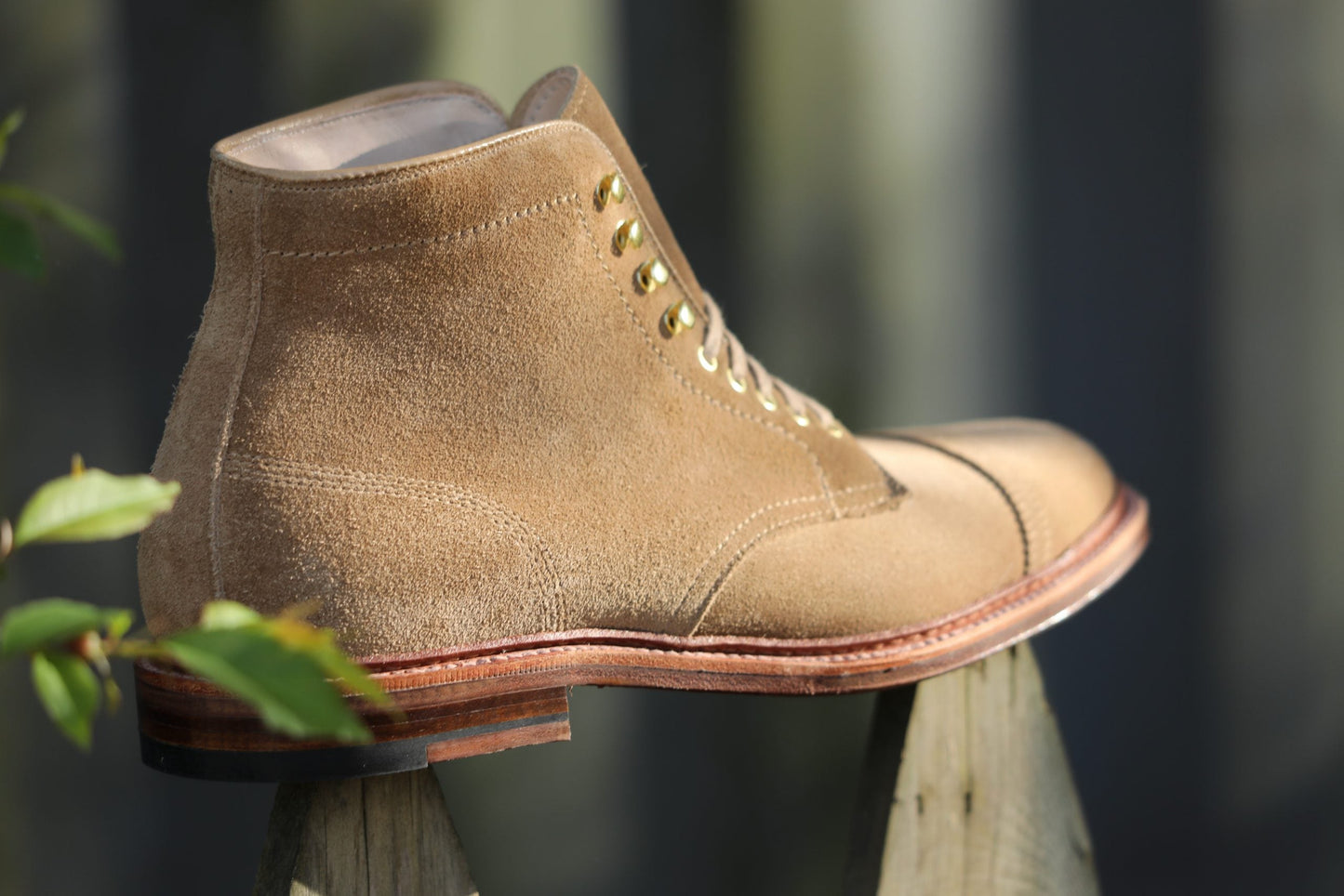 The Alden Madison D2685H - 2/2 Cap Toe Boot in Tan Suede, featuring golden eyelets and a brown wooden sole, sits on a wooden fence post with blurred greenery in the background, elegantly embodying Barrie Lasts design essence.
