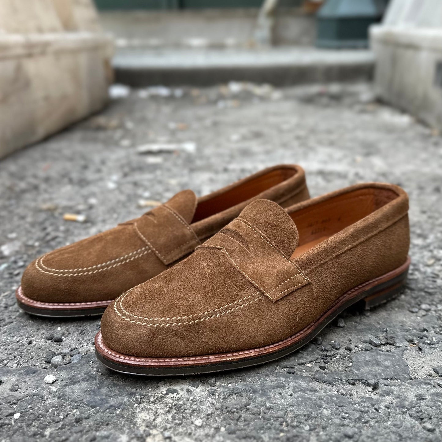 The Alden Madison 6221L Unlined Penny Mocc in Snuff Suede features a classic brown suede design with light stitching and mocc toe, shown on a textured concrete surface. It has a smooth finish and rubber sole against a blurred urban background.
