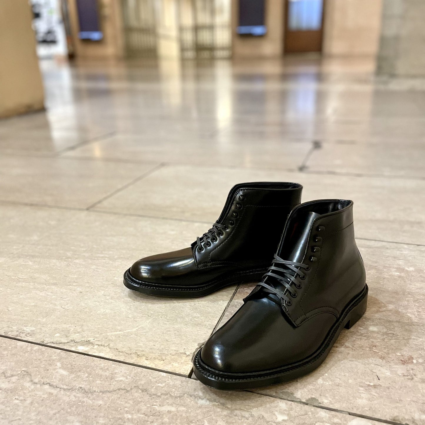 A pair of Alden Madison’s D6842H - Plain Toe Director Boot in Black Shell Cordovan sits on a light-colored tiled floor. The background displays an elegant interior with blurred columns and doorways, suggesting a spacious setting.