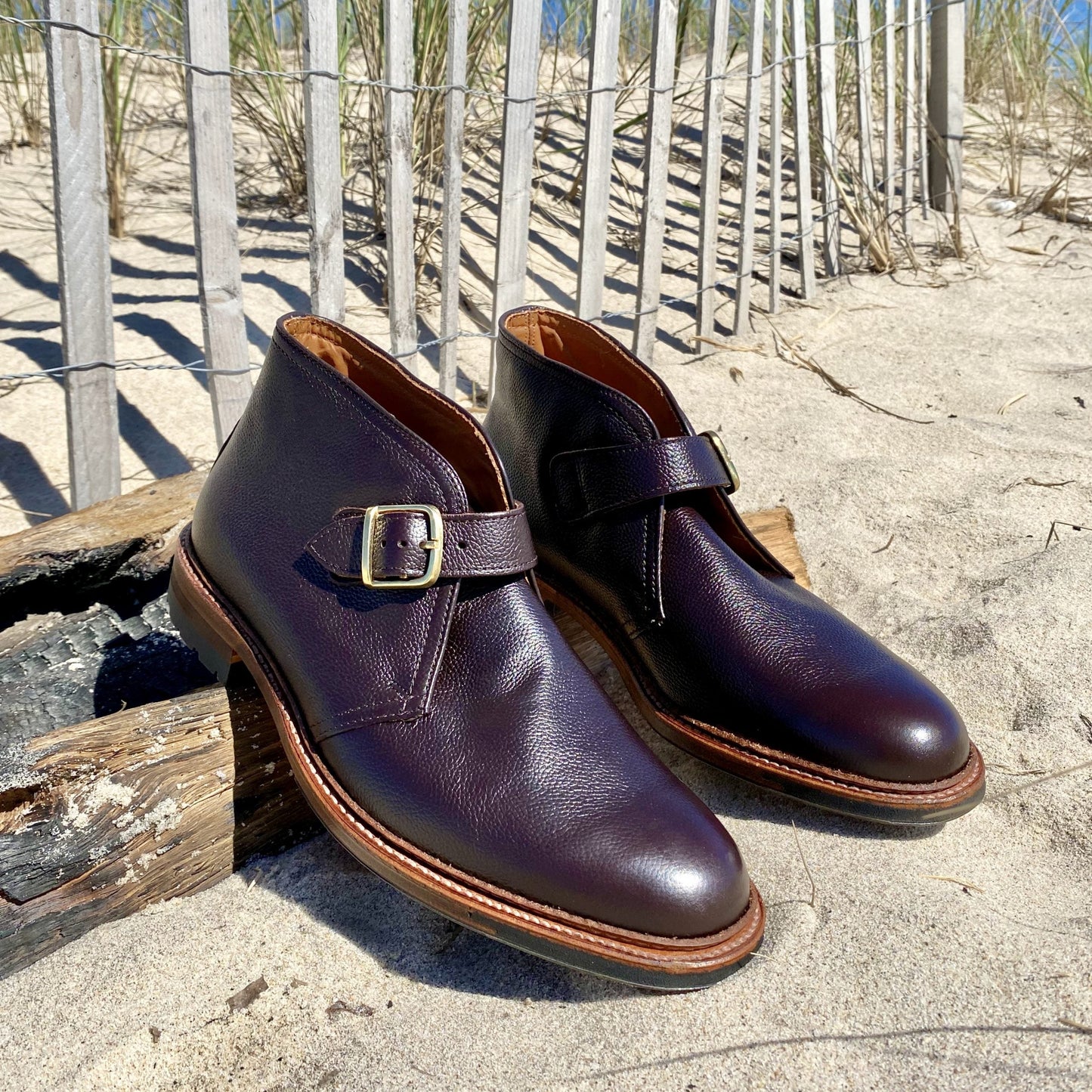 A pair of Alden Madisons D9704C - George Boots in Brown Regina Grain rest on driftwood in the sand, with a wooden fence and dune grass against a clear blue sky in the background.