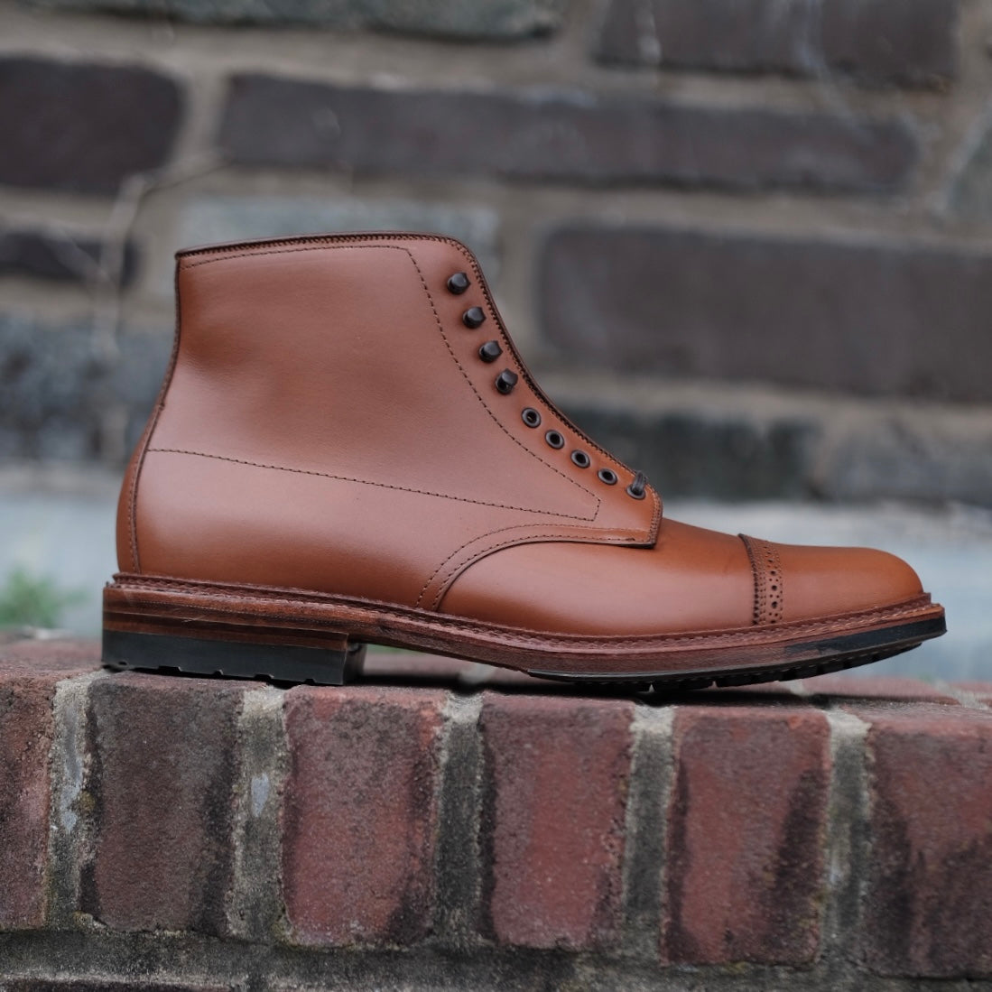 An Alden Madison D2825HC Jumper Boot in burnished tan calfskin rests on a brick ledge against a blurred wall. It features black eyelets, a smooth upper, slightly textured commando sole, and intricate stitching on the cap toe and sides.