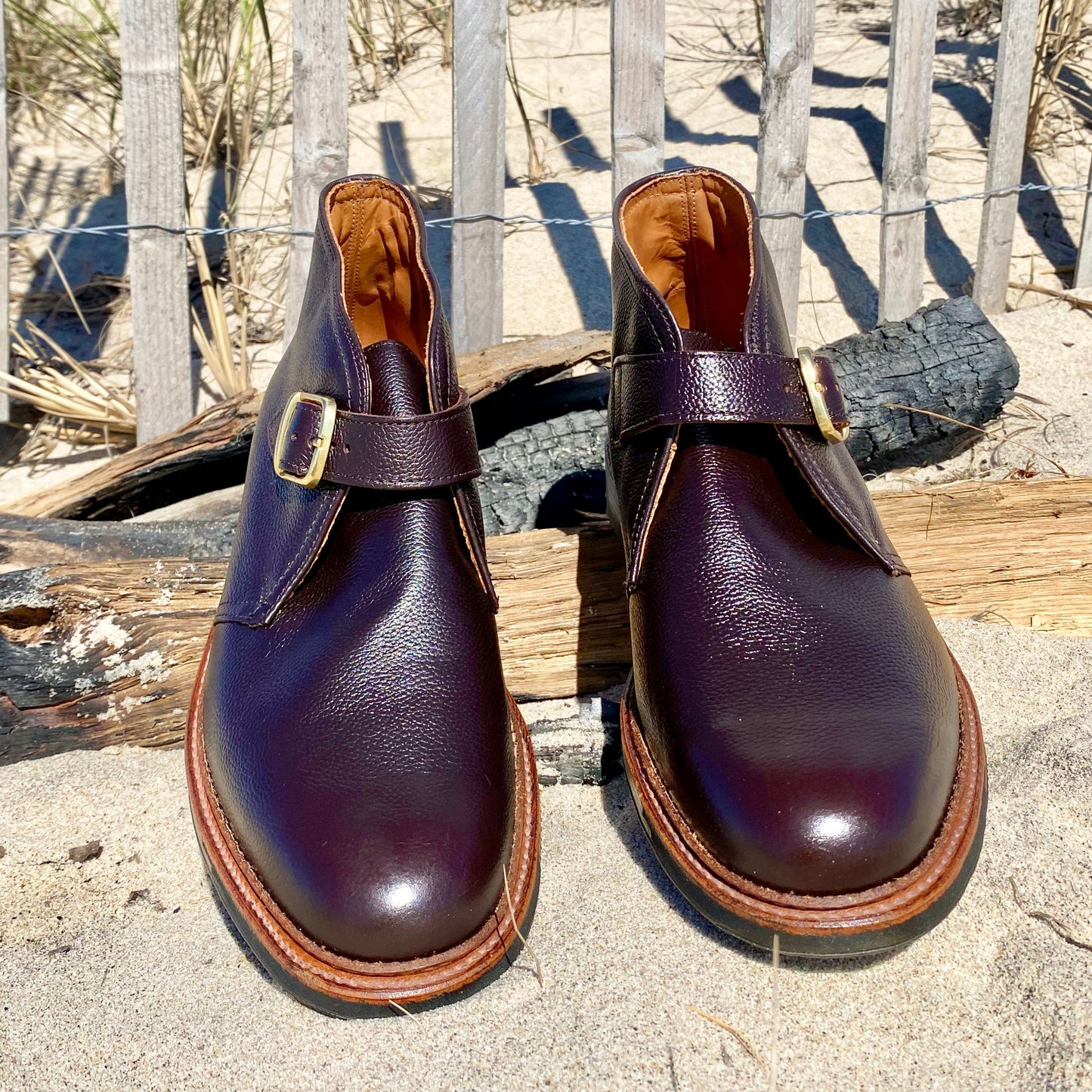 The Alden Madison D9704C - George Boot in Brown Regina Grain rests on sandy ground near weathered wood planks and a rustic fence, with dry grass visible in the background.