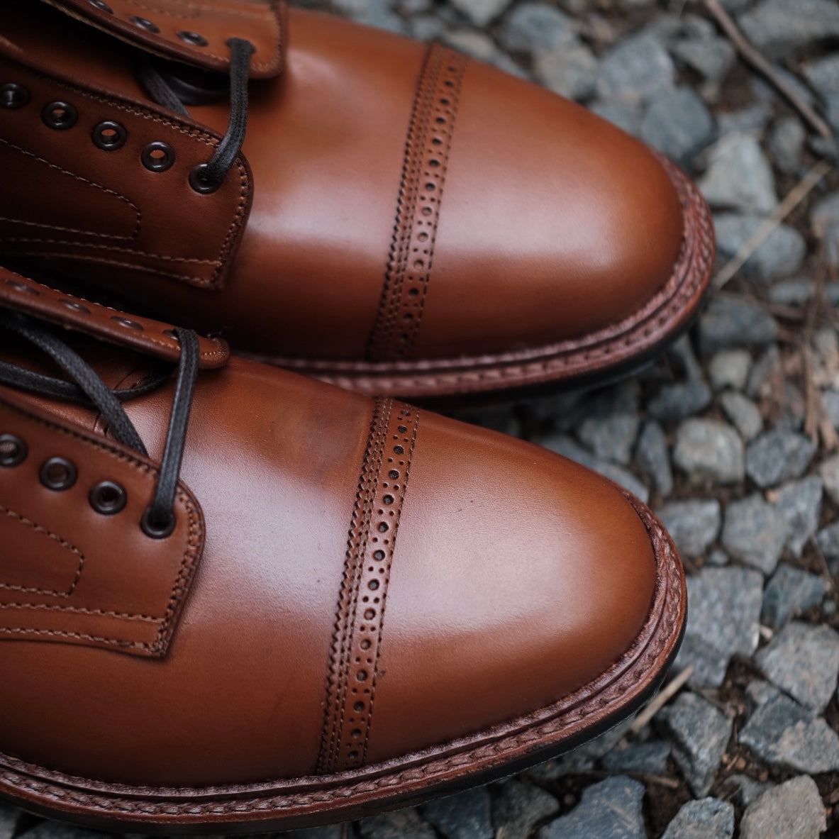 Close-up of Alden Madisons D2825HC - Jumper Boot in burnished tan calfskin, crafted with decorative toe cap perforations and dark brown laces, elegantly placed on a cobblestone surface.