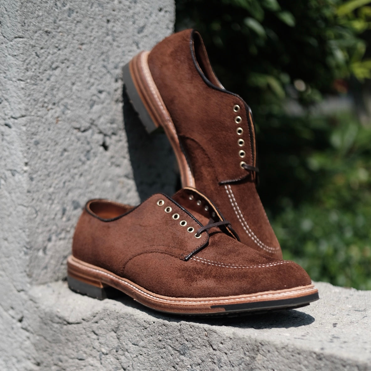 Alden Madisons D2604C Indy Shoes in Tobacco Reverse Chamois, crafted on the Trubalance Last, feature light stitching and laces. One shoe stands upright on concrete with greenery while the other leans against it.