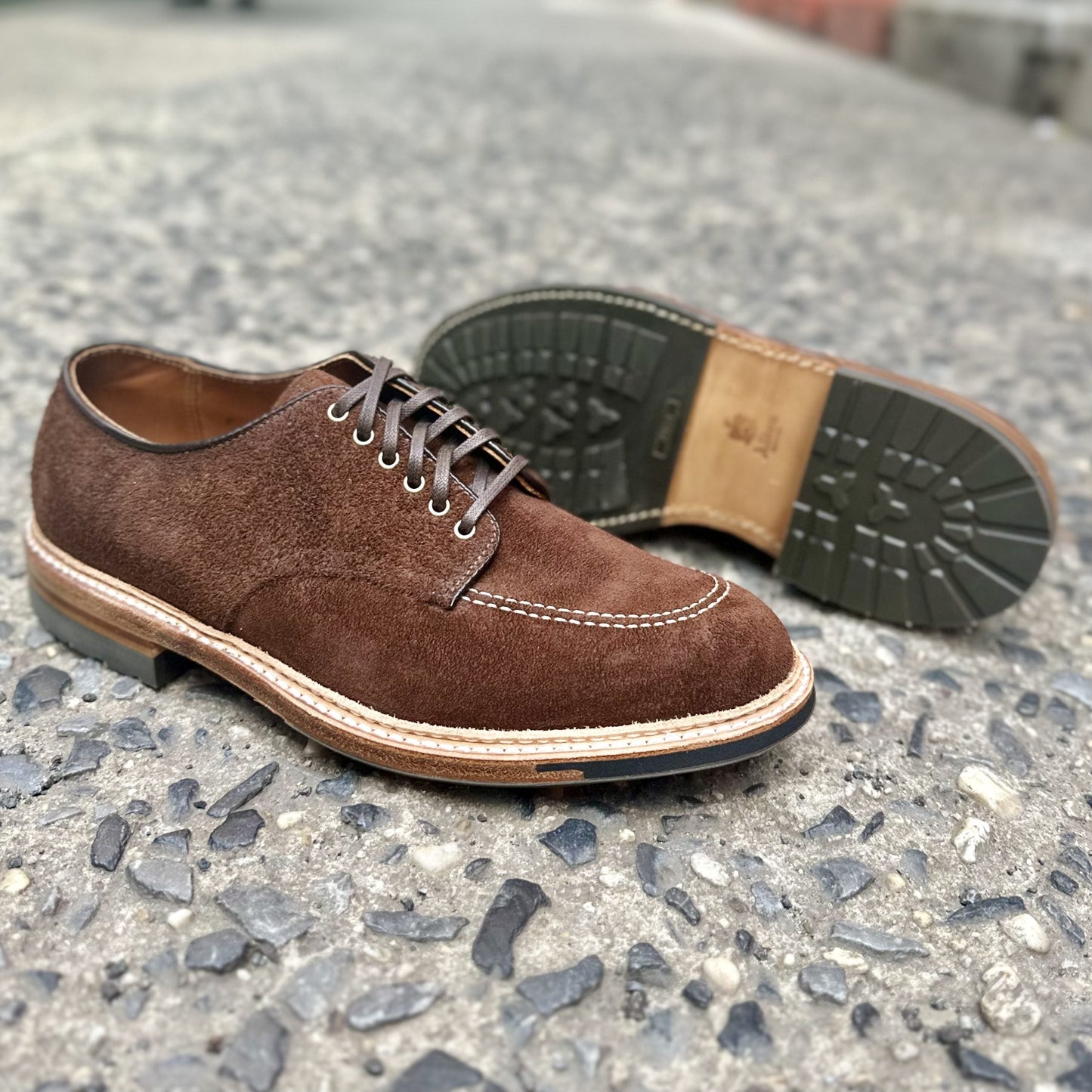 A pair of Alden Madisons D2604C - Indy Shoes in Tobacco Reverse Chamois rests on a rough concrete surface. The front shoe stands upright, displaying its side and laces; the rear shoe is on its side, revealing the Trubalance Last sole with intricate stitching and tread pattern.