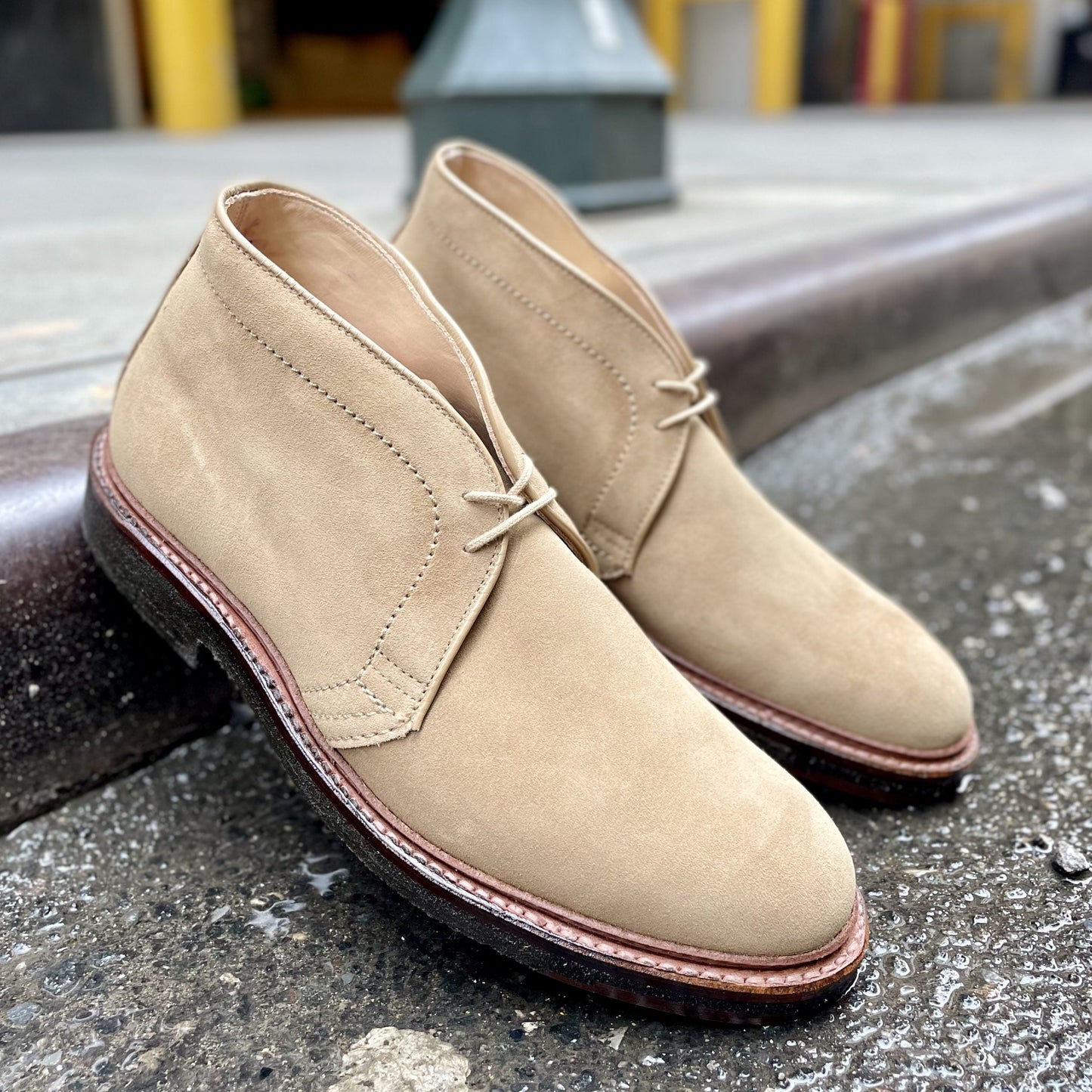 A pair of Alden Madisons 14580 - Desert Chukka Boot in Tan Suede with brown soles and laces rests against a curb on a paved street. The Barrie Last design is prominent, set against blurred outdoor elements like a lamppost and colorful buildings.