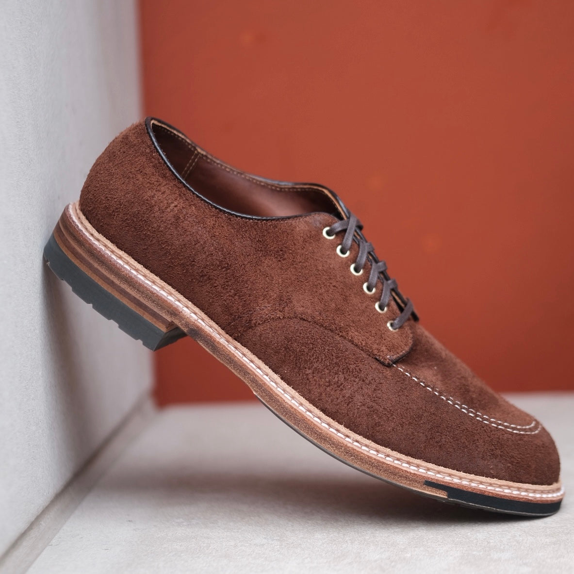The Alden Madison D2604C Indy Shoe, crafted from Tobacco Reverse Chamois, is a brown suede dress shoe with dark laces and a thick stitched sole. Highlighted by the Trubalance Last, it is shown on a white surface against a warm rust-colored background.