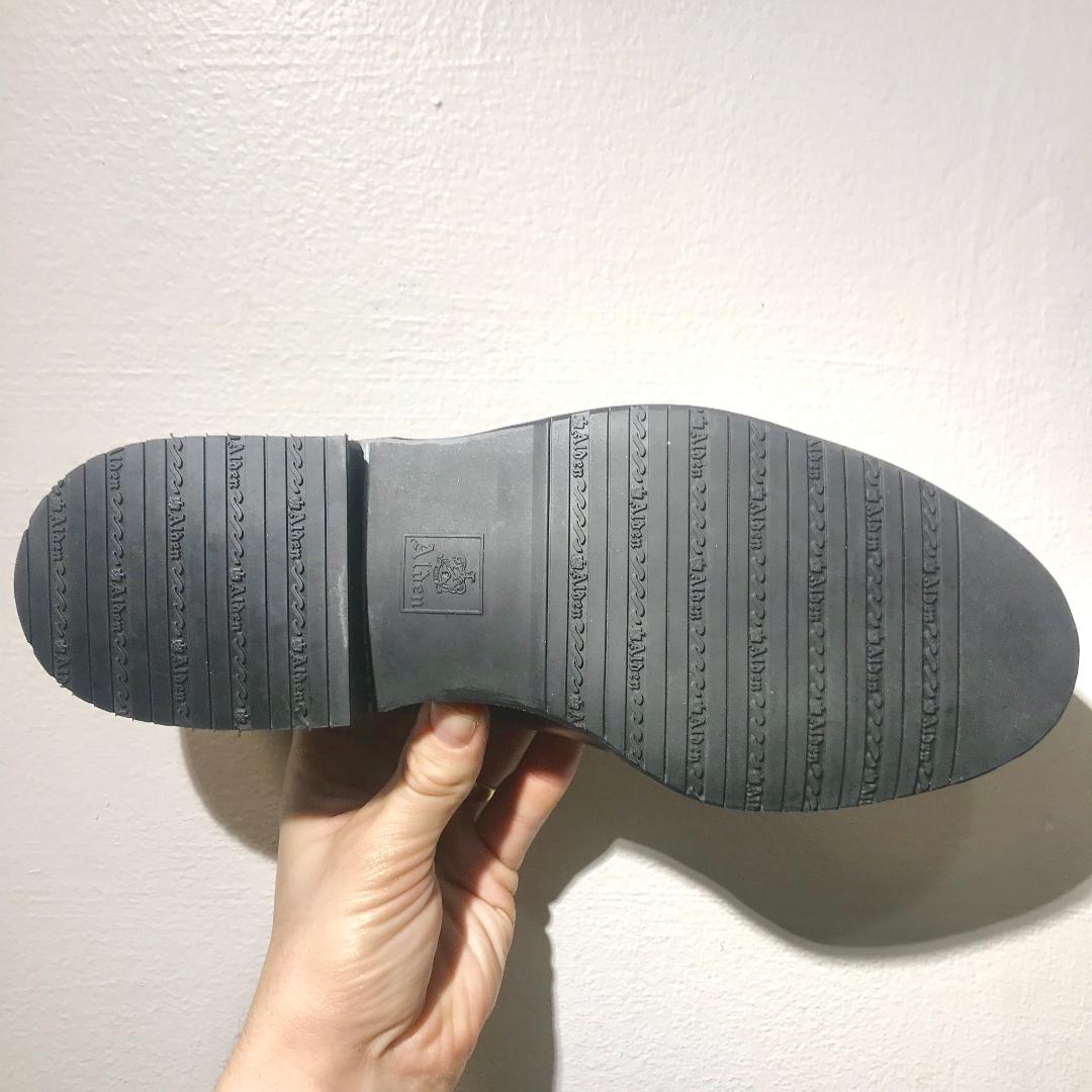A hand displays the sole of the Alden Madison 702 - Mocc Toe Blucher in Snuff Suede against a white background, showcasing its sleek black rubber design with parallel textured grooves and a central logo imprint, perfect for dress shoes.