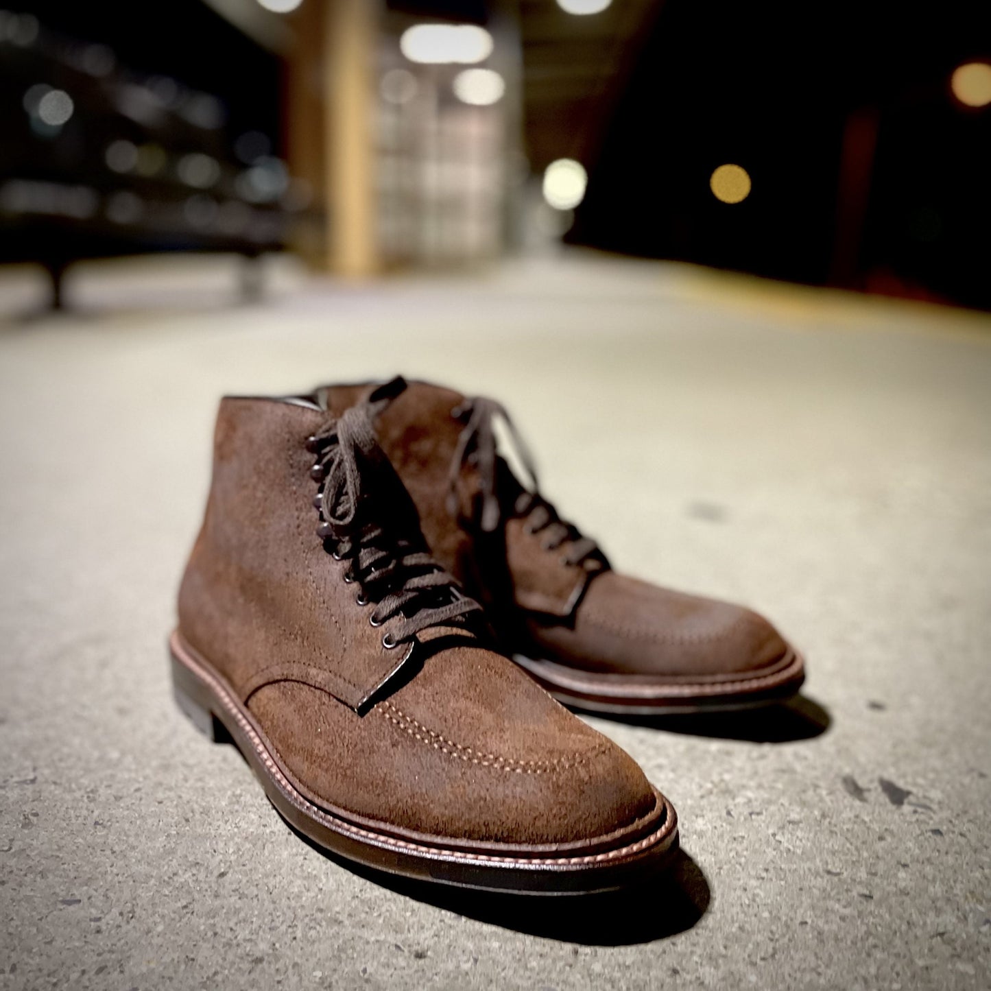 A pair of Alden Madisons 4015HC - Indy Boots in Tobacco Reverse Chamois with dark laces is on a concrete floor. The background is blurred, featuring dim lighting with soft yellow hues and bokeh effects.