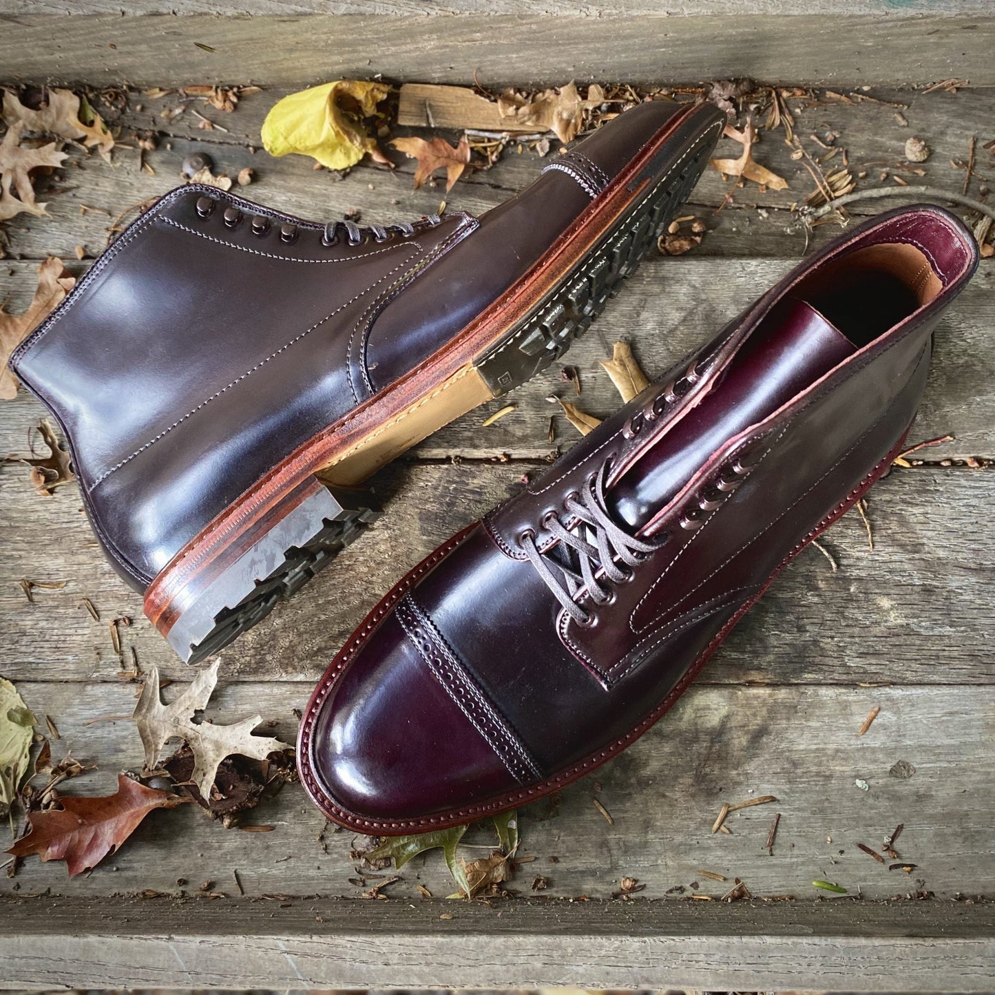 A pair of Alden Madisons D6861HC - Jumper Boots in Color 8 Shell Cordovan with a Commando Sole rest on a wooden surface, surrounded by autumn leaves. The neatly done Speed Hook Lacing highlights their classic, sturdy design.