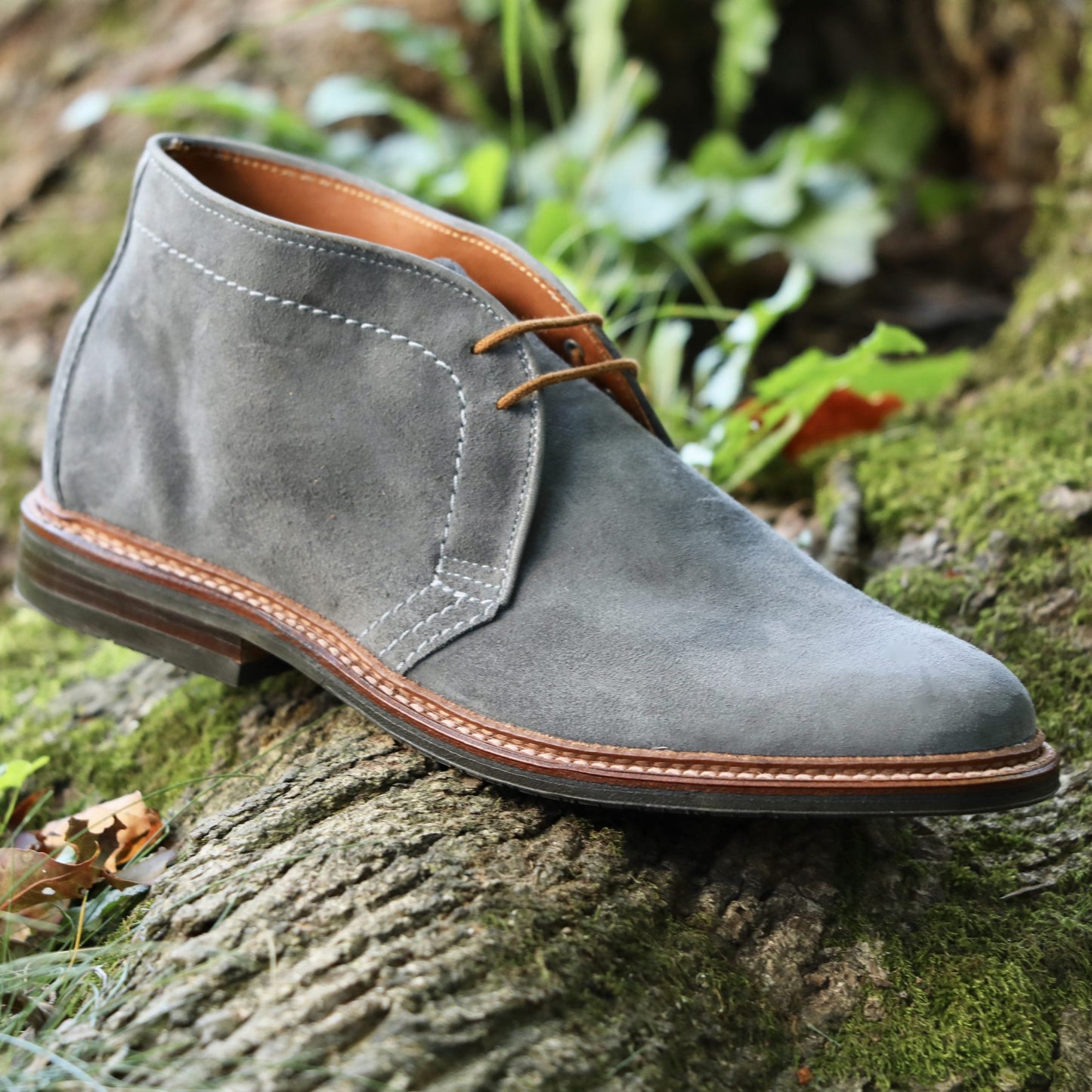 The Alden Madison 1592L Chukka Boot with L3 Sole in Lead Suede, featuring brown laces and a Reverse Antique Welt, sits atop a mossy tree trunk outdoors amid lush green foliage.