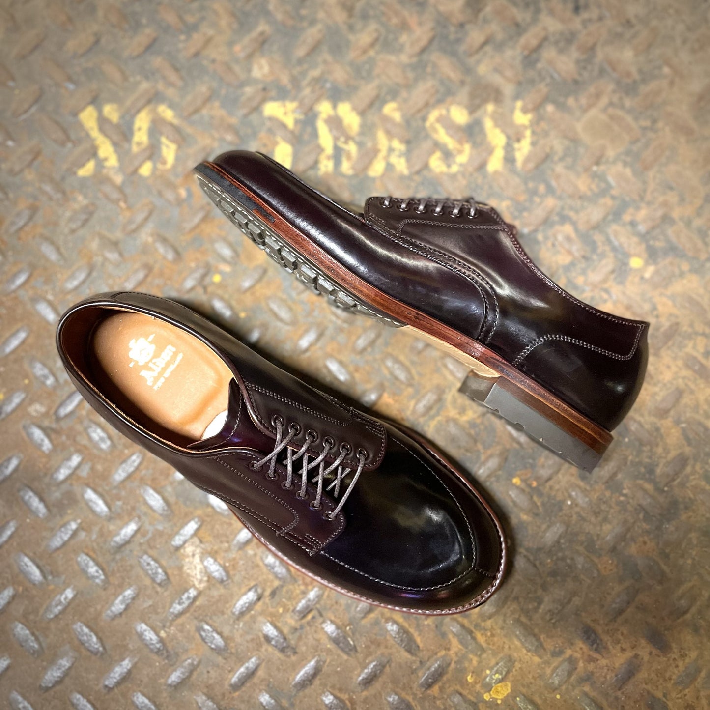 A glossy pair of Alden Madisons D1629C - V Tip Split Toe Blucher in Color 8 Shell sits on a textured metal surface; one shoe is upright and laced, while the other is on its side, showcasing its rugged Commando Sole with visible brand markings.