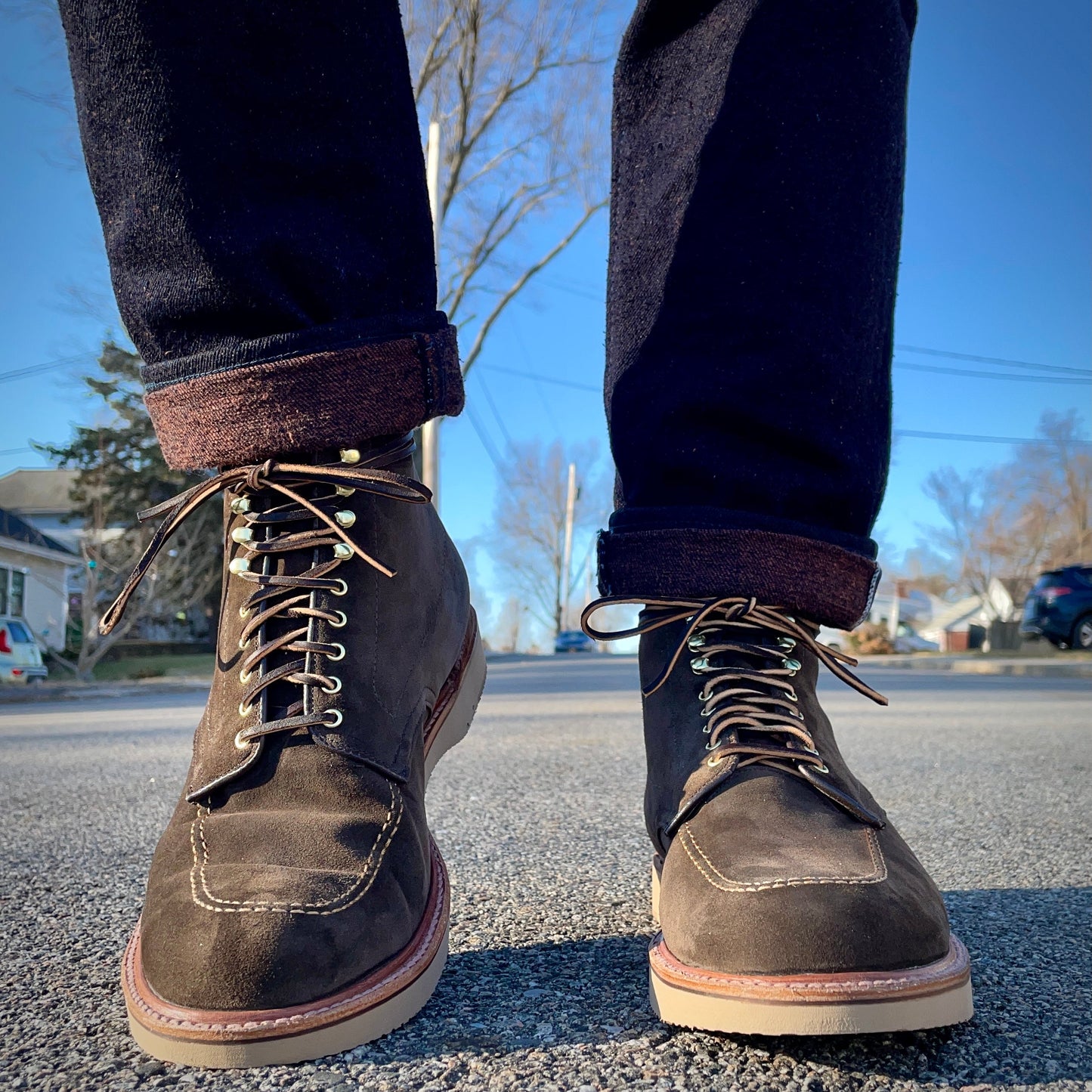 Close-up of a person wearing Alden Madisons Duncan Indy Boot in Loden Suede, standing on a pavement. The jeans are cuffed, revealing details crafted on a Truebalance Last. A clear blue sky, trees, and part of a residential area with houses form the background.