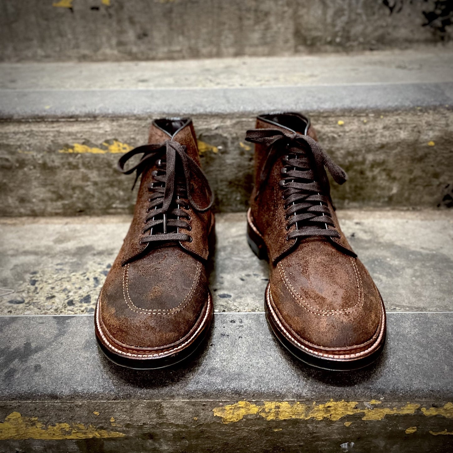 A pair of Alden Madisons 4015HC - Indy Boot in Tobacco Reverse Chamois with laces rests on concrete steps. Crafted from reverse chamois leather, the boots display a textured, rustic look with visible stitching. The steps show wear, with yellow paint chipping off.