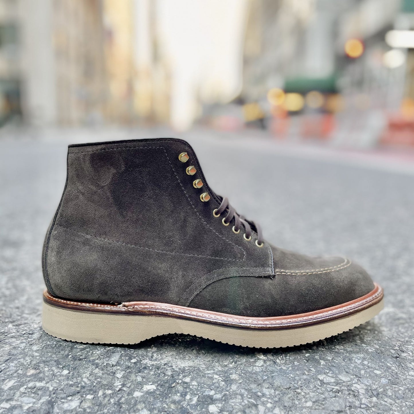 The Alden Madison D0938H - Duncan Indy Boot in Loden Suede, featuring a storm welt, light tan sole, and brass eyelets, is shown on a city street with blurred buildings and sidewalks in the background.