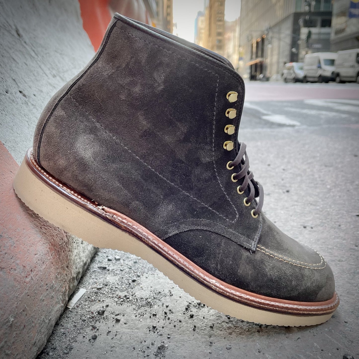 The Alden Madison D0938H - Duncan Indy Boot in Loden Suede, featuring golden eyelets and a beige sole, rests against a concrete surface. The background showcases an urban street with buildings, vehicles, and orange construction barriers.