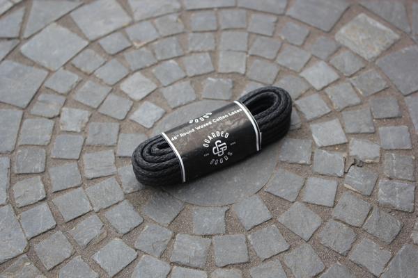 A pair of 48-inch round boot laces from Alden Madison is coiled and elegantly displayed on a circular mosaic of gray stone tiles, nestled in their sleek branded packaging.