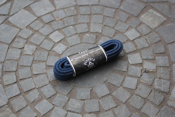A coiled set of Alden Madisons Round Boot Laces - 48 Inch, in navy blue, sits at the center of a stone mosaic with concentric circles. The black and white label contrasts the textured pattern. Perfect length for any footwear.