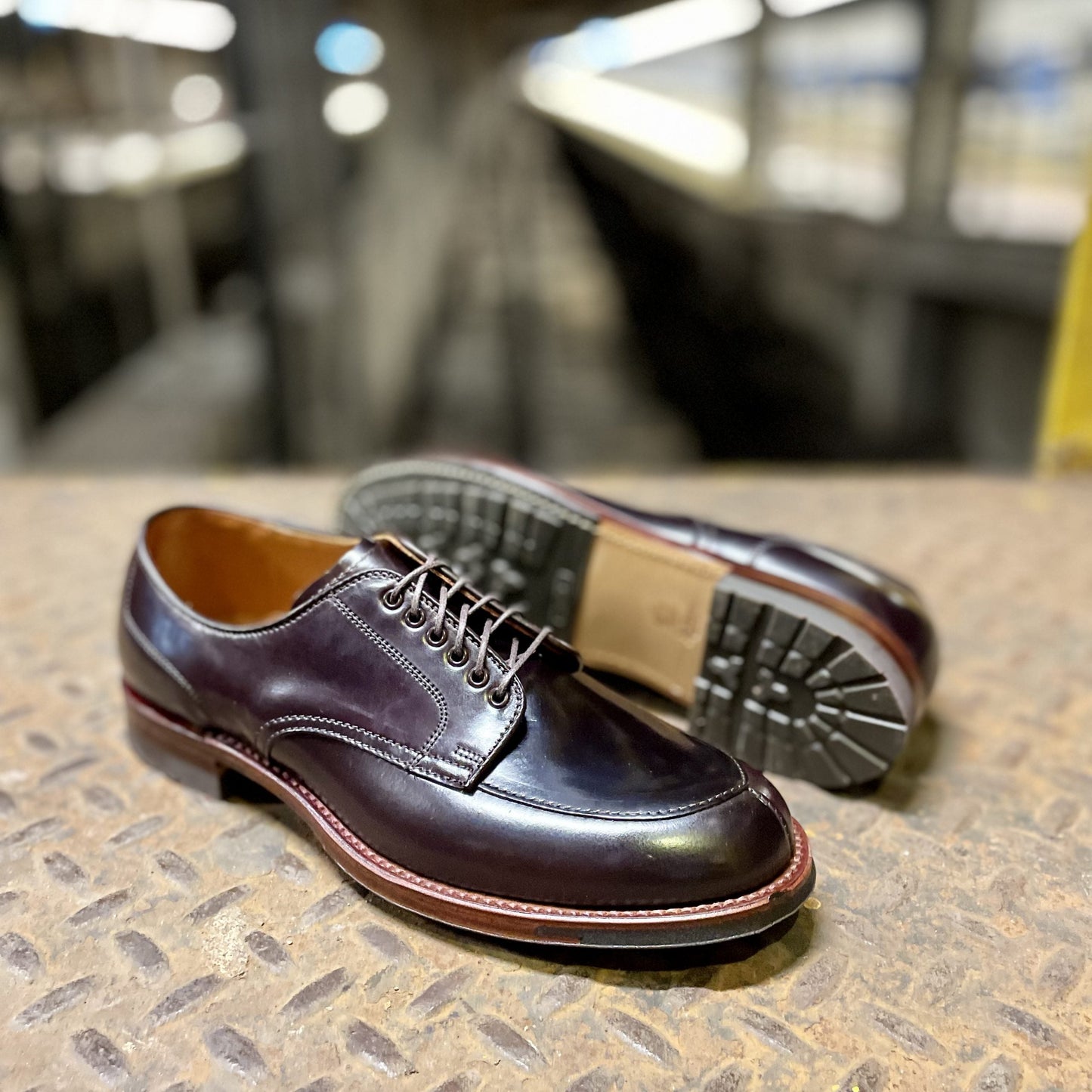 A pair of Alden Madisons D1629C - V Tip Split Toe Bluchers in Color 8 Shell rests on a textured metal surface. One shoe lies flat, the other shows off its sturdy commando sole. A blurred industrial background completes the scene.