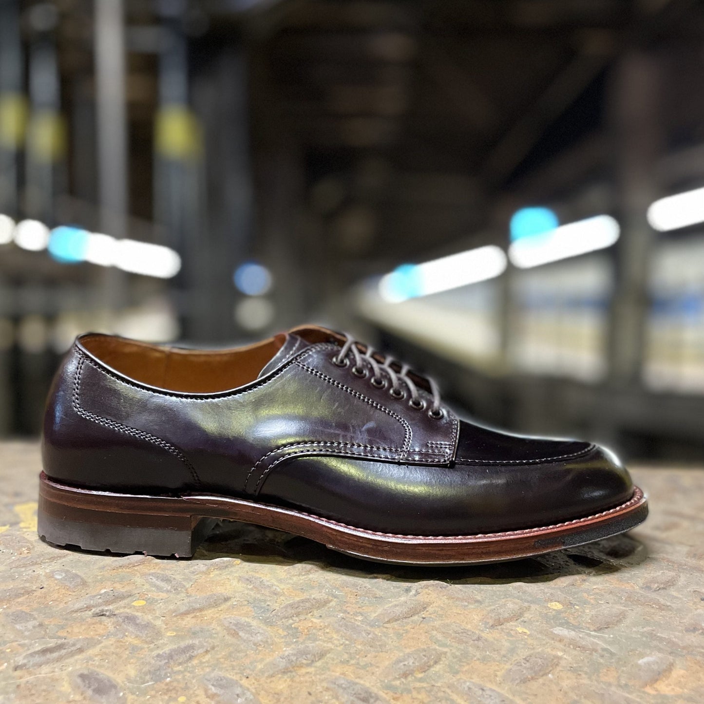 On a textured surface, the Alden Madison D1629C - V Tip Split Toe Blucher in Color 8 Shell is showcased with its polished black leather, Commando Sole, and brown trim. Industrial lighting enhances the shoes sleek design and craftsmanship against a blurred background.