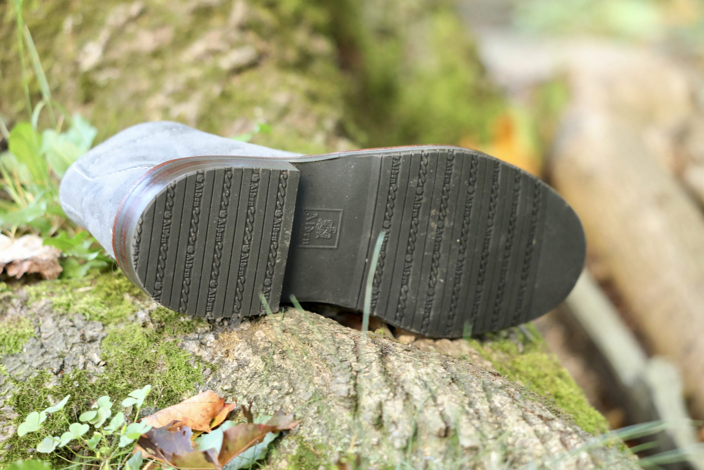 A close-up of the L3 Sole on the Alden Madison 1592L Chukka Boot in Lead Suede, resting on a mossy tree trunk amid green grass and fallen leaves in an outdoor setting.