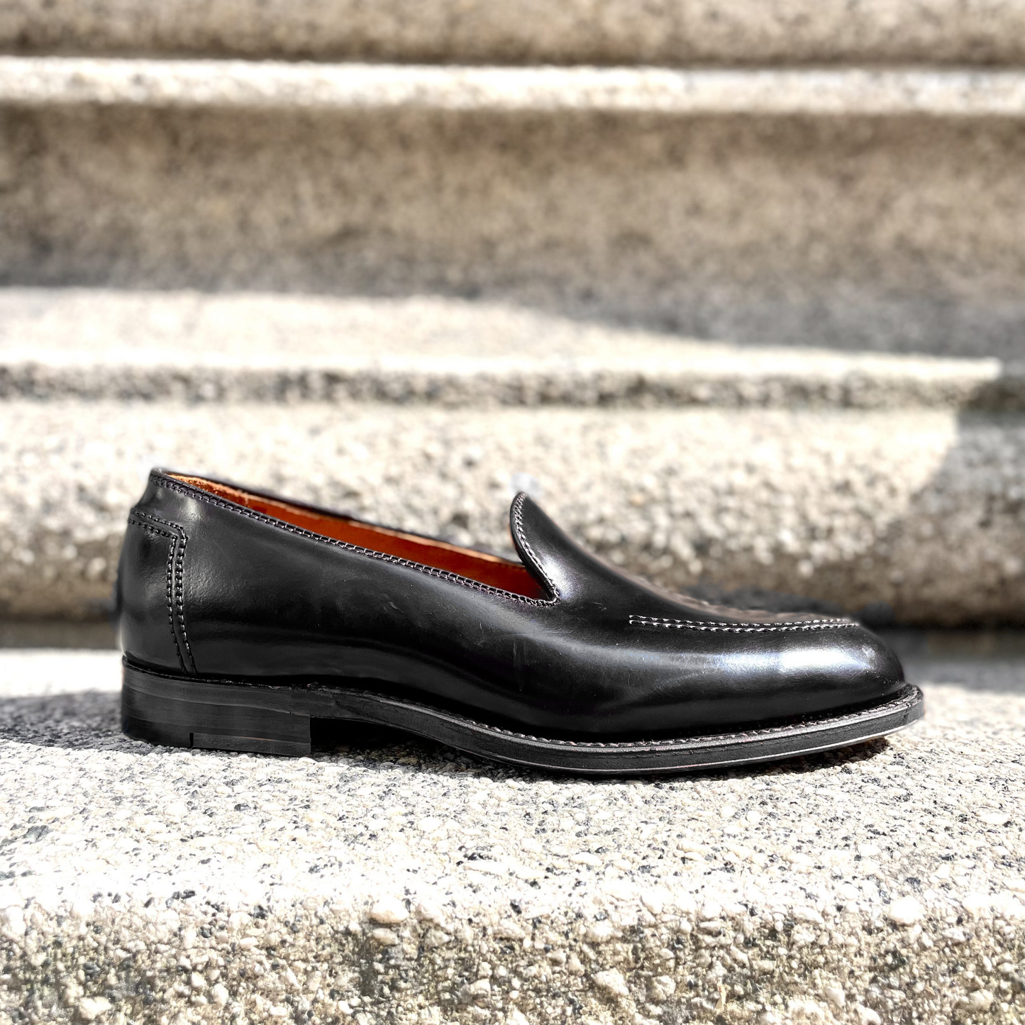 The Alden Madison D2117 - Vanderbilt Slip On in Black Shell Cordovan rests on textured stone steps, showcasing a sleek design with Brooks Heel Foxing and detailed stitching. Its polished surface gleams beautifully in the sunlight.