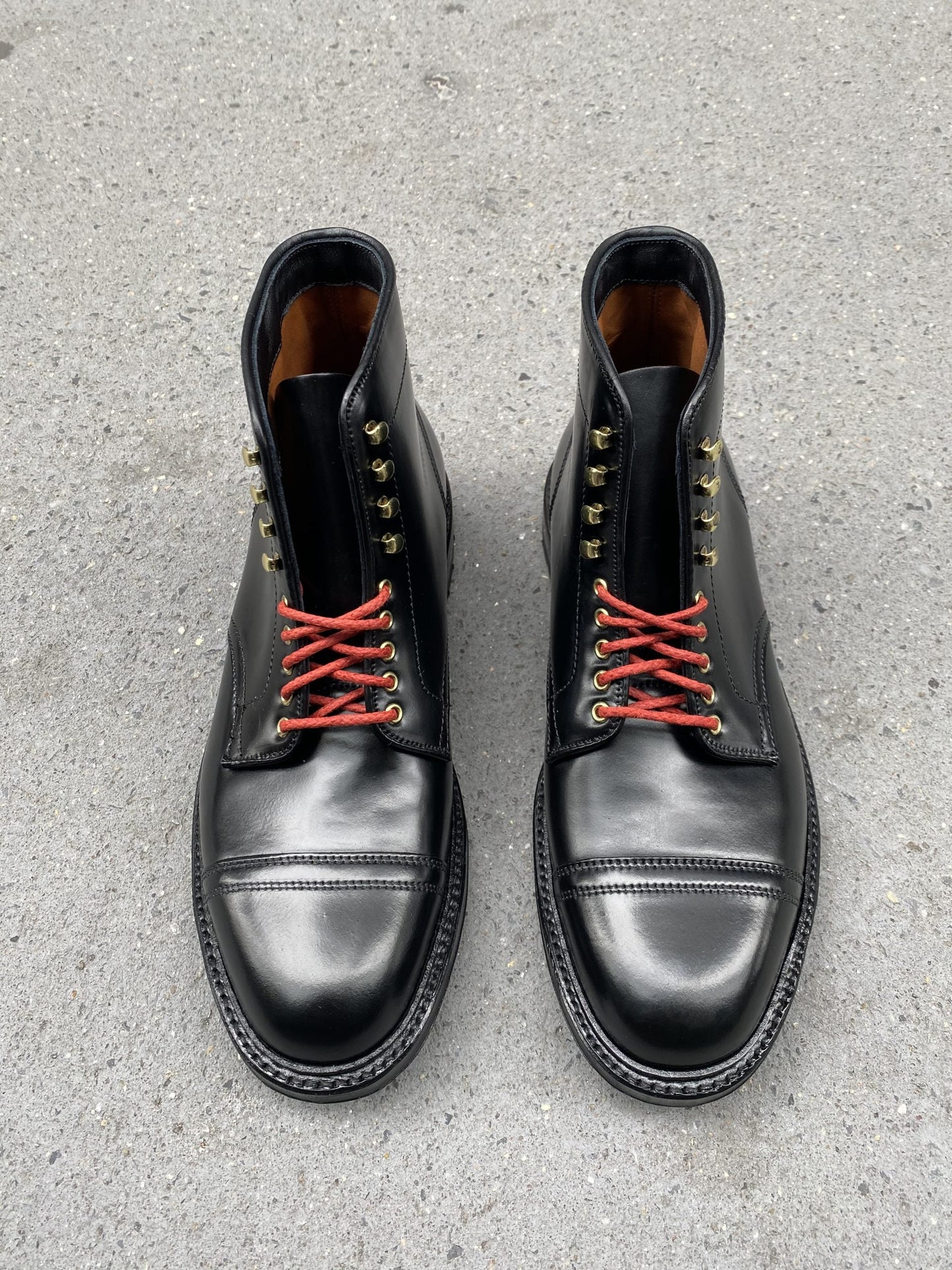 A pair of Alden Madisons D9867HC- Stitchup 2/2 Boots in Black Shell Cordovan with red laces, gold eyelets, and a commando sole sits on concrete, capturing the Stitchdown.com style.