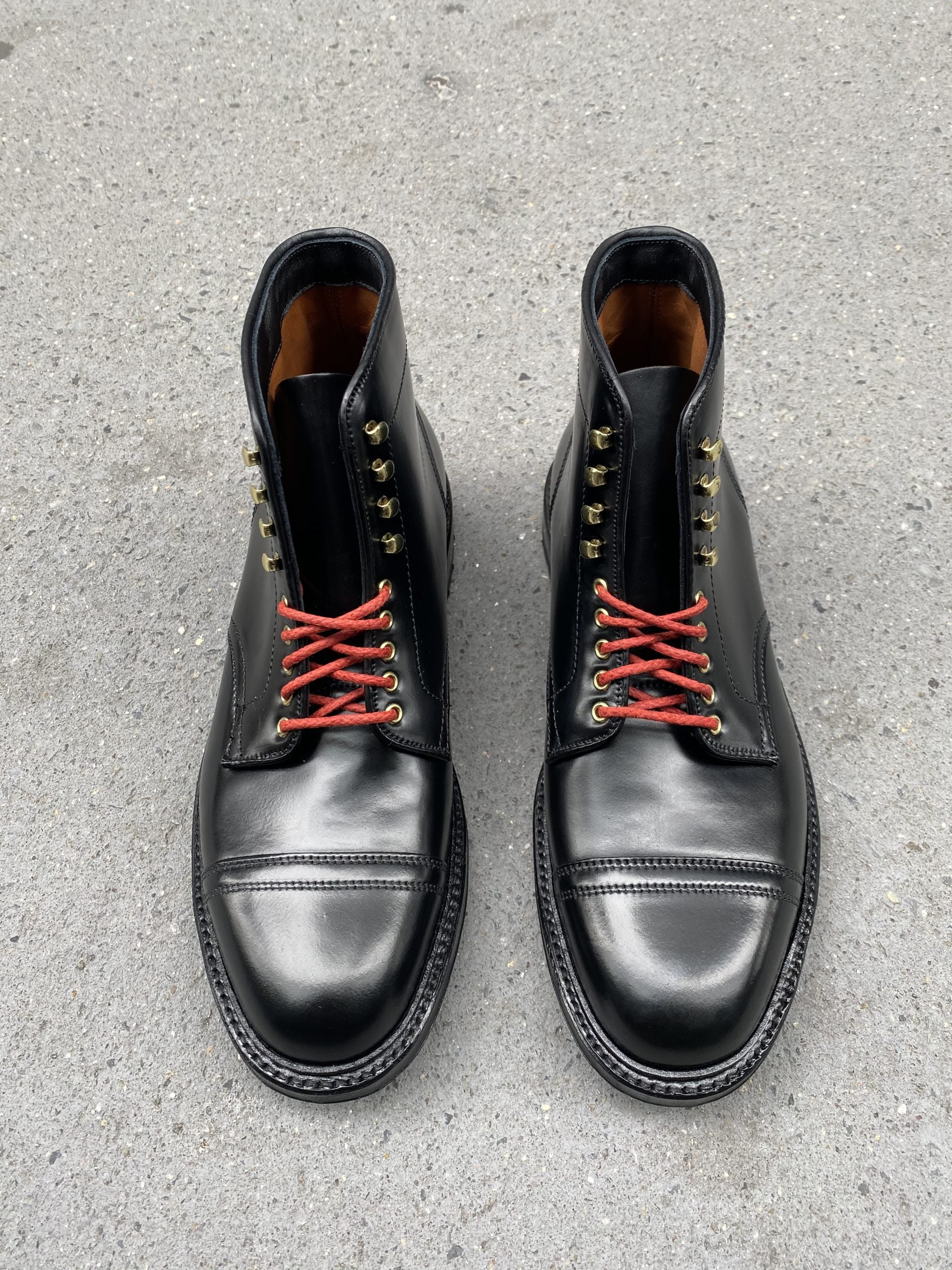 A pair of Alden Madisons D9867HC- Stitchup 2/2 Boots in Black Shell Cordovan with red laces, gold eyelets, and a commando sole sits on concrete, capturing the Stitchdown.com style.