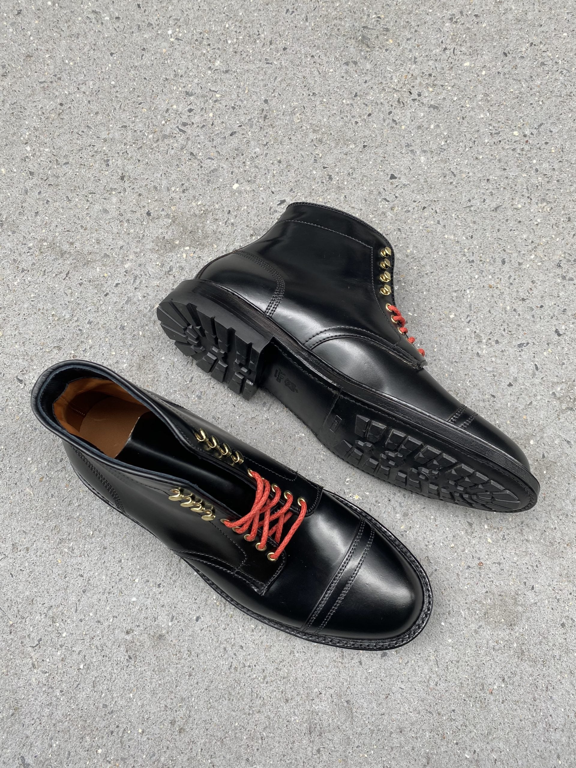 Alden Madisons D9867HC- Stitchup 2/2 Boot in Black Shell Cordovan, featuring black leather and red laces, is displayed on a gray concrete surface. One boot lies on its side highlighting the Commando Sole with deep treads, while the other stands upright.