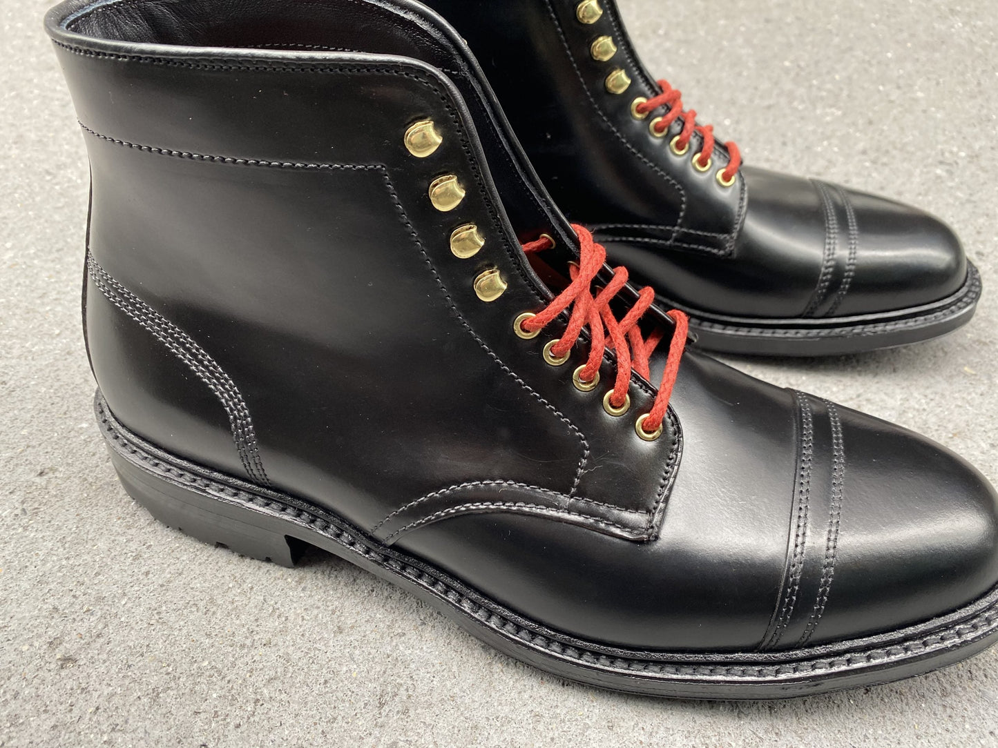 A pair of Alden Madison D9867HC- Stitchup 2/2 boots in black shell cordovan with red laces and gold eyelets showcased on a gray surface, featuring detailed stitching, a cap toe design, and a sturdy commando sole from Stitchdown.com.