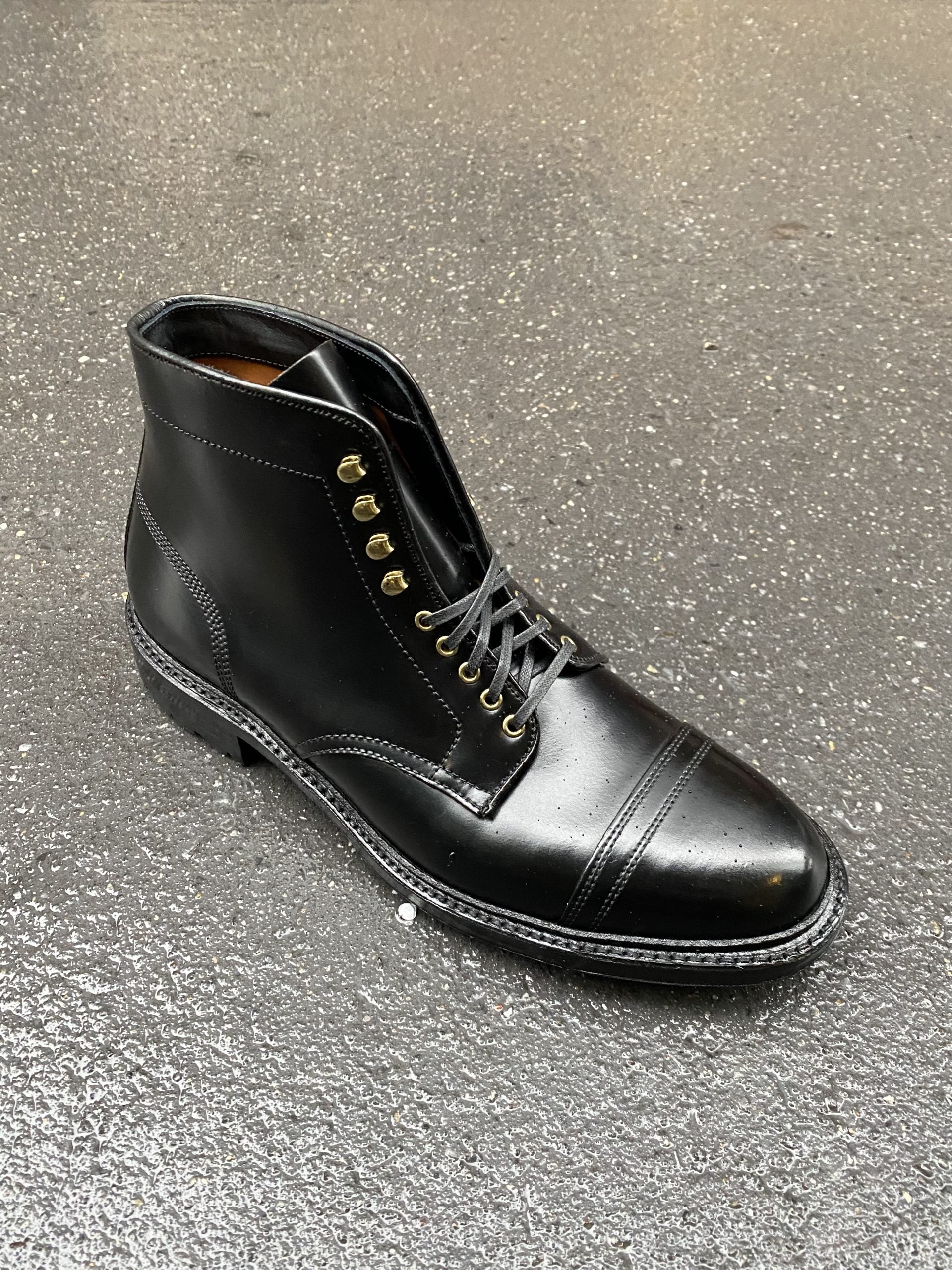 A single Alden Madison D9867HC- Stitchup 2/2 Boot in Black Shell Cordovan, with metallic eyelets and a rugged Commando Sole, rests on wet gray pavement, highlighting its polished finish and detailed stitching.