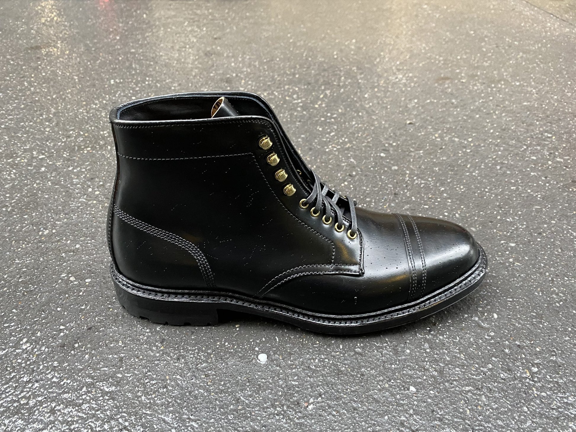 Positioned on a textured gray surface, the Alden Madison D9867HC-Stitchup 2/2 Boot in sleek Black Shell Cordovan features laces, metal eyelets, and a polished finish. The boots solid stitching showcases durability and style on its Commando Sole.