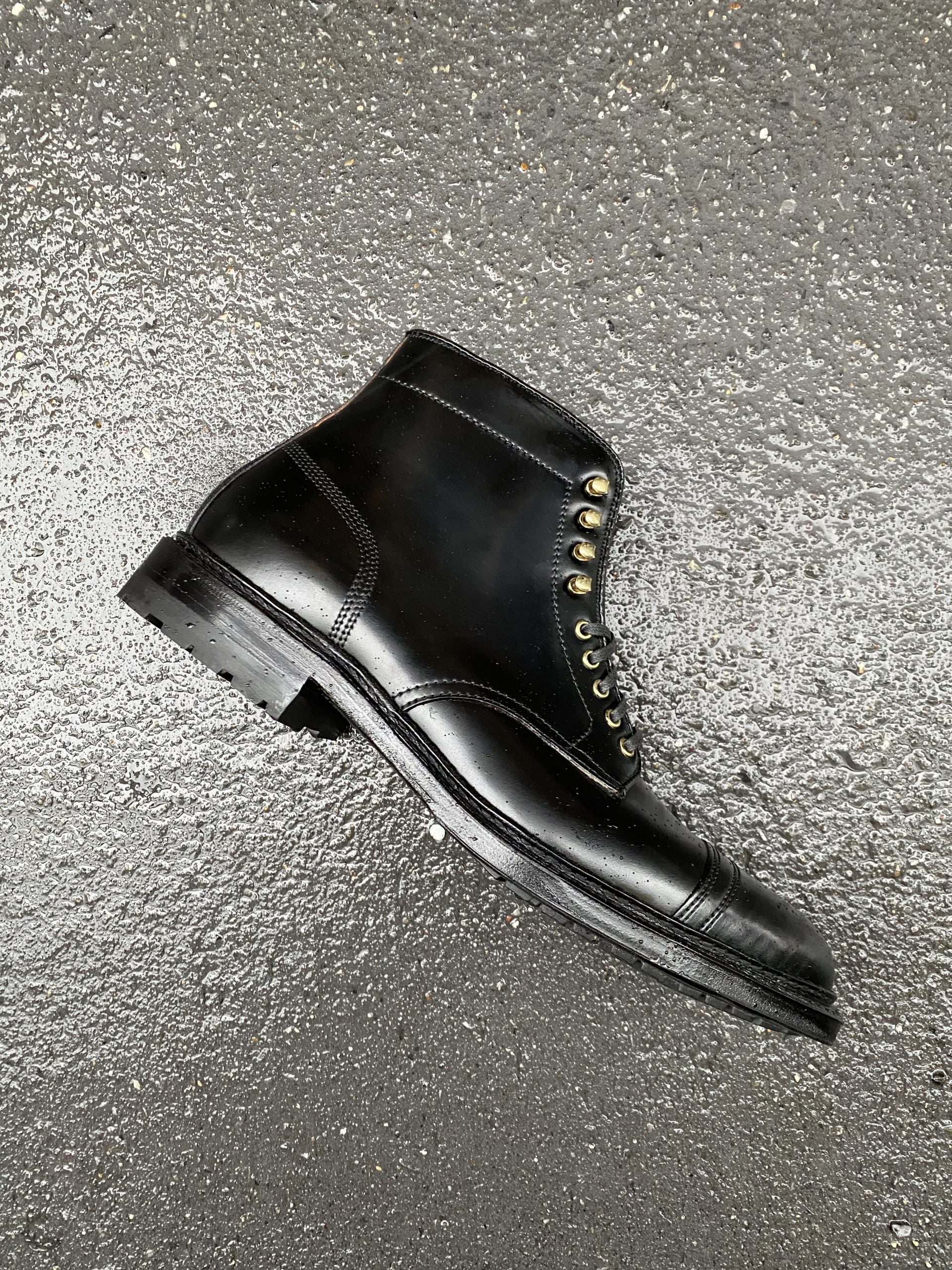 A single D9867HC-Stitchup 2/2 Boot in Black Shell Cordovan by Alden Madison, featuring brass eyelets, a Commando Sole, cap toe, and visible stitching, lies on a speckled gray surface.